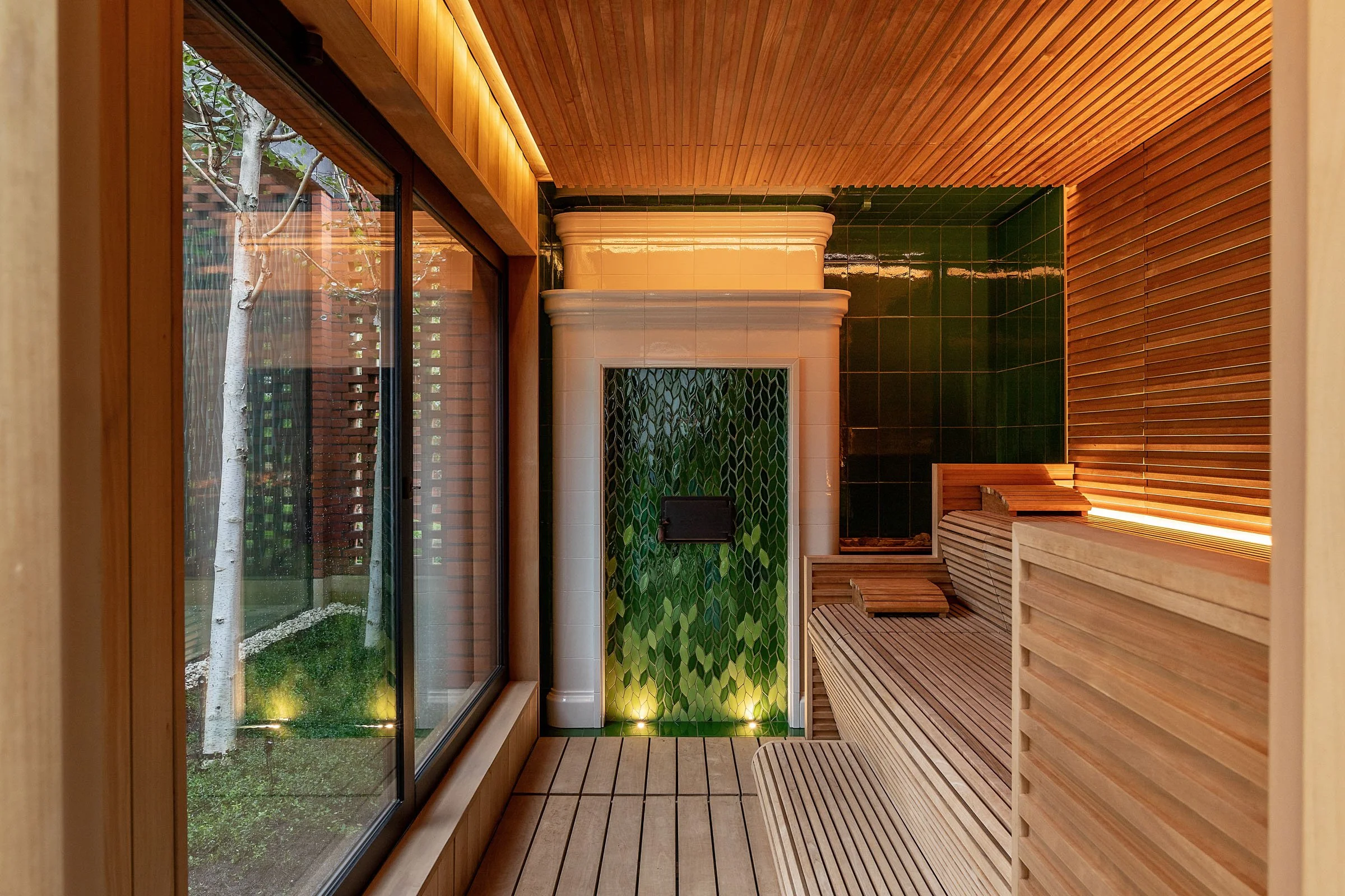 Interior view of a modern sauna room with wooden benches, green tiled wall, glass window showing outside trees, and warm ambient lighting.