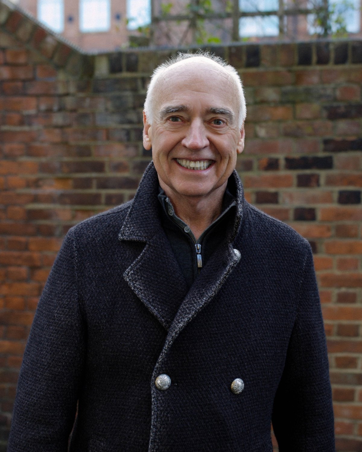 A middle-aged man with light skin, receding gray hair, and wearing a dark patterned shirt with rolled-up sleeves, standing against a blue background.