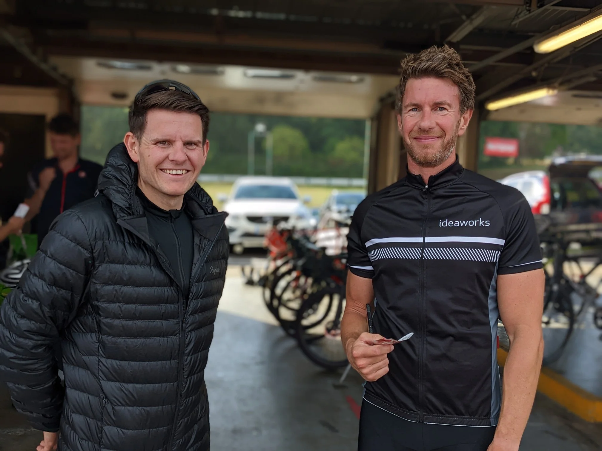 Two men in cycling gear standing under a covered area, smiling, with bicycles and cars in the background.