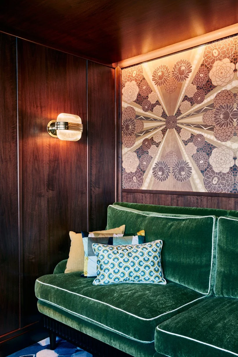 Interior living room corner with a dark wood paneled wall, a gold wall sconce, a green velvet sofa with patterned and solid pillows, and a floral patterned wall art.