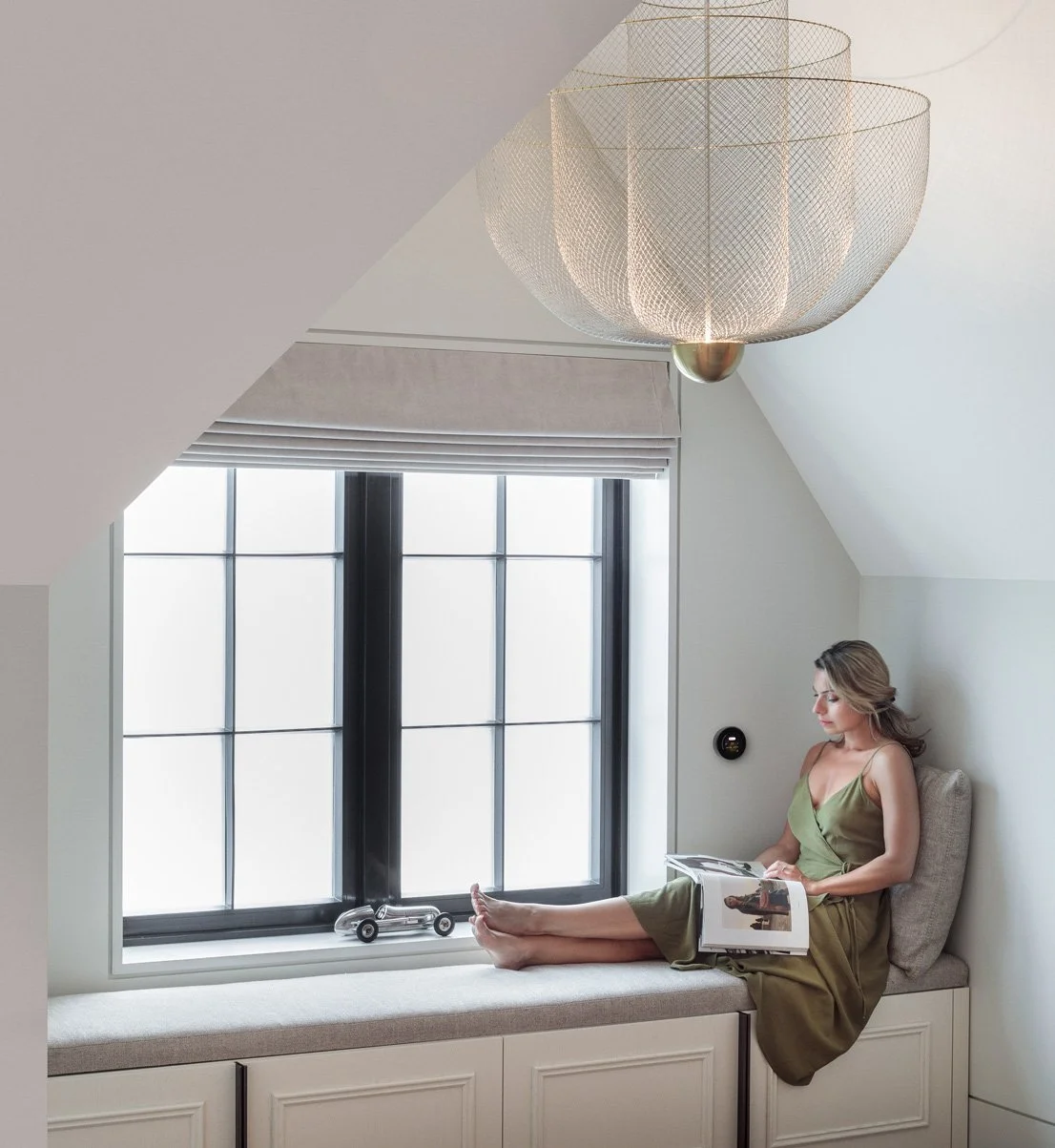 A woman in a green dress is sitting on a window seat in a luxury home, reading a magazine with a small toy car on the sill, inside a bright room with a large window and a decorative ceiling light. There is a Turn 2 home control device on the wall.