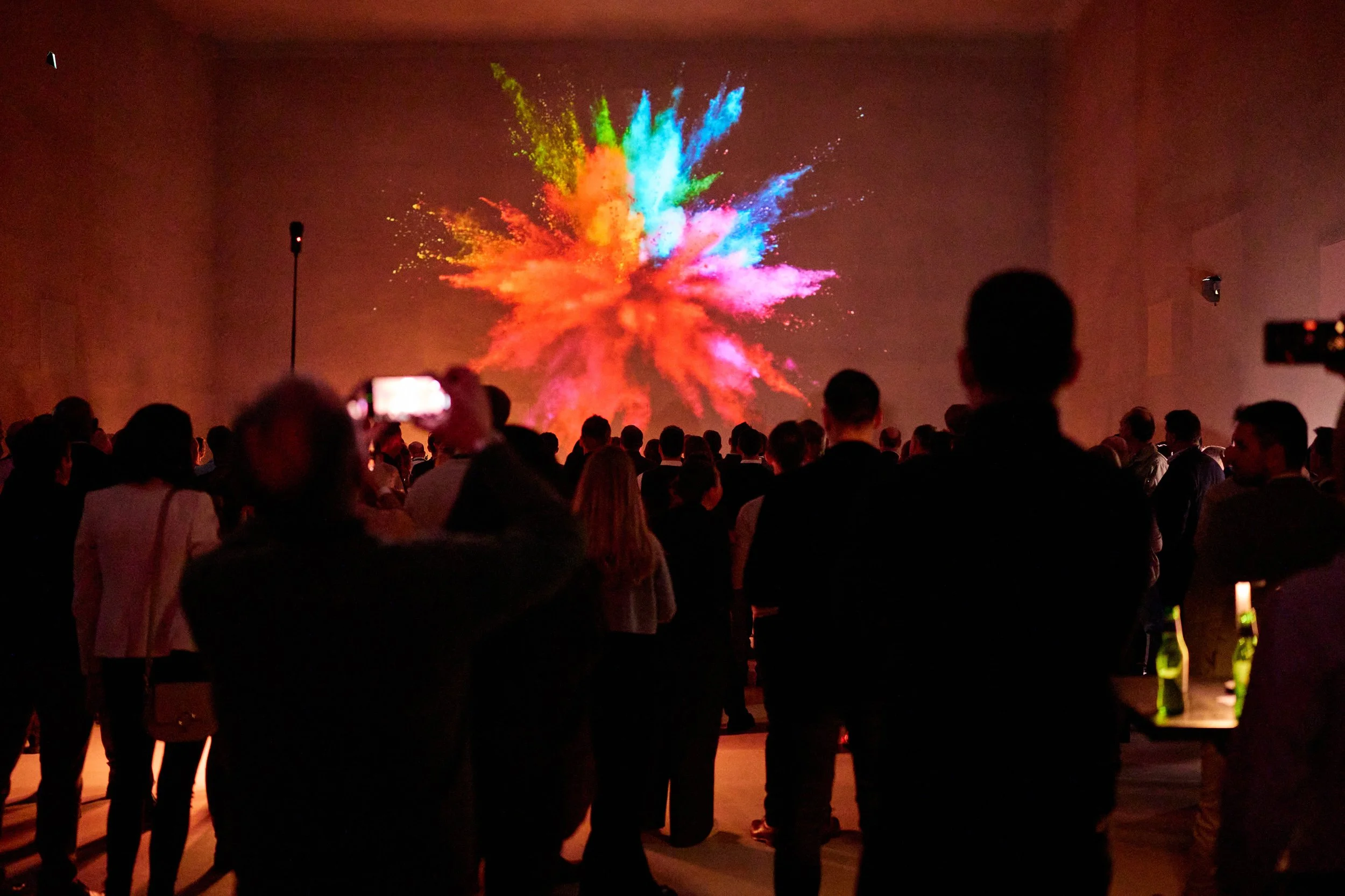 A large group of people watching a colorful fireworks display in a dark indoor setting.