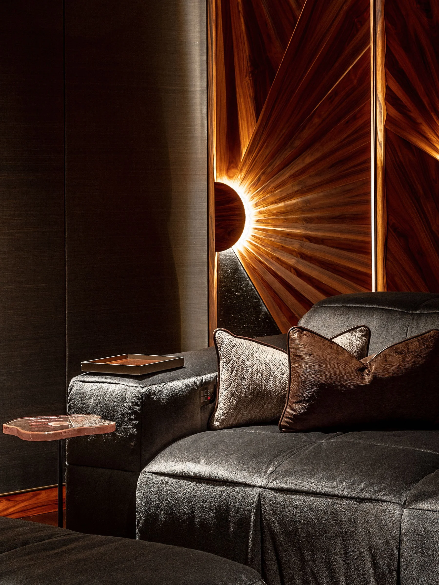 Interior living room with a dark velvet sofa, gold and brown cushions, a side table with a tray, and a wooden wall with a sunburst design illuminated by a glowing sun and black sphere.