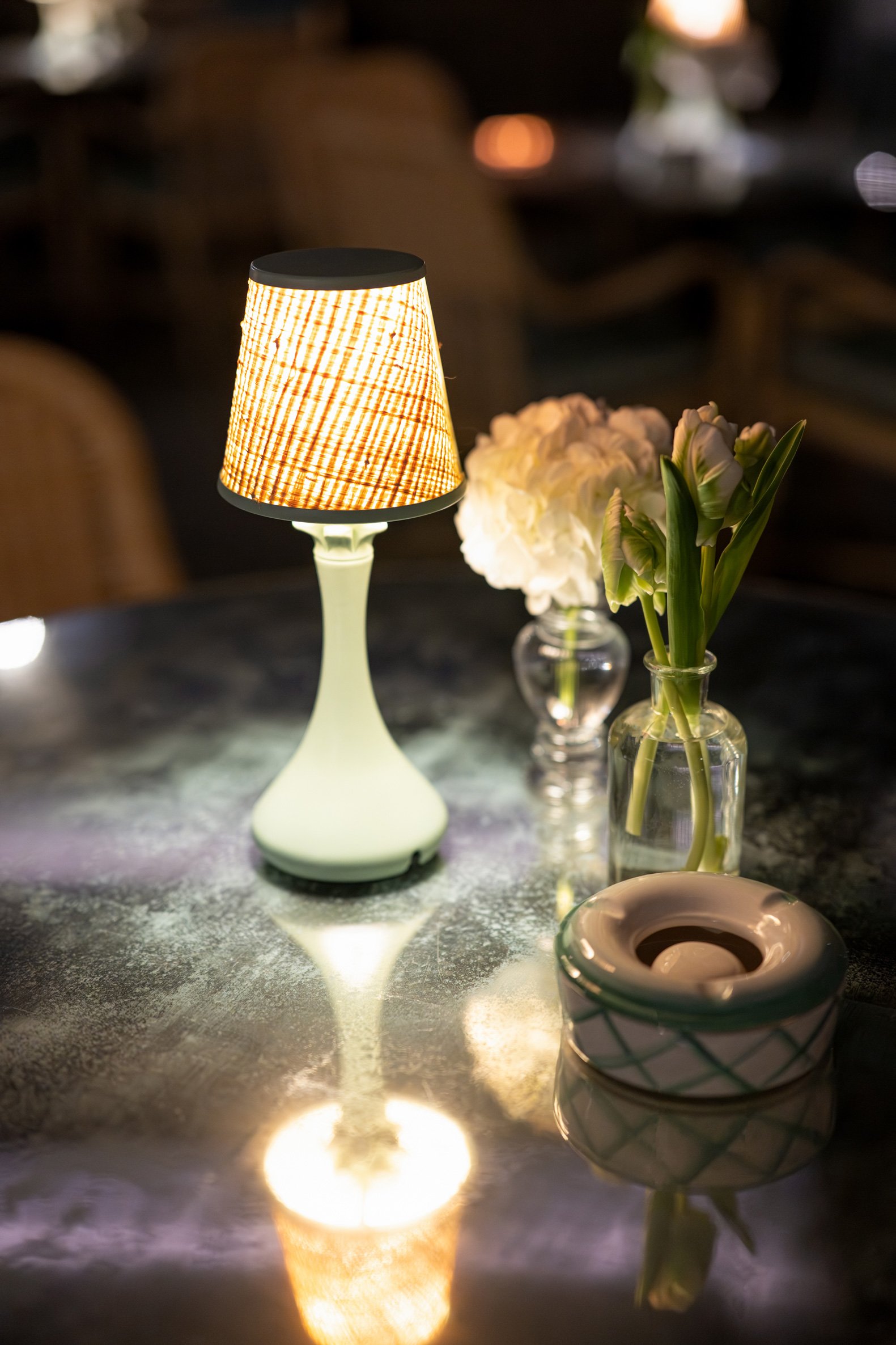 A table with a small lamp, a glass vase with white flowers, and a decorative ceramic container, illuminated in a cozy setting.