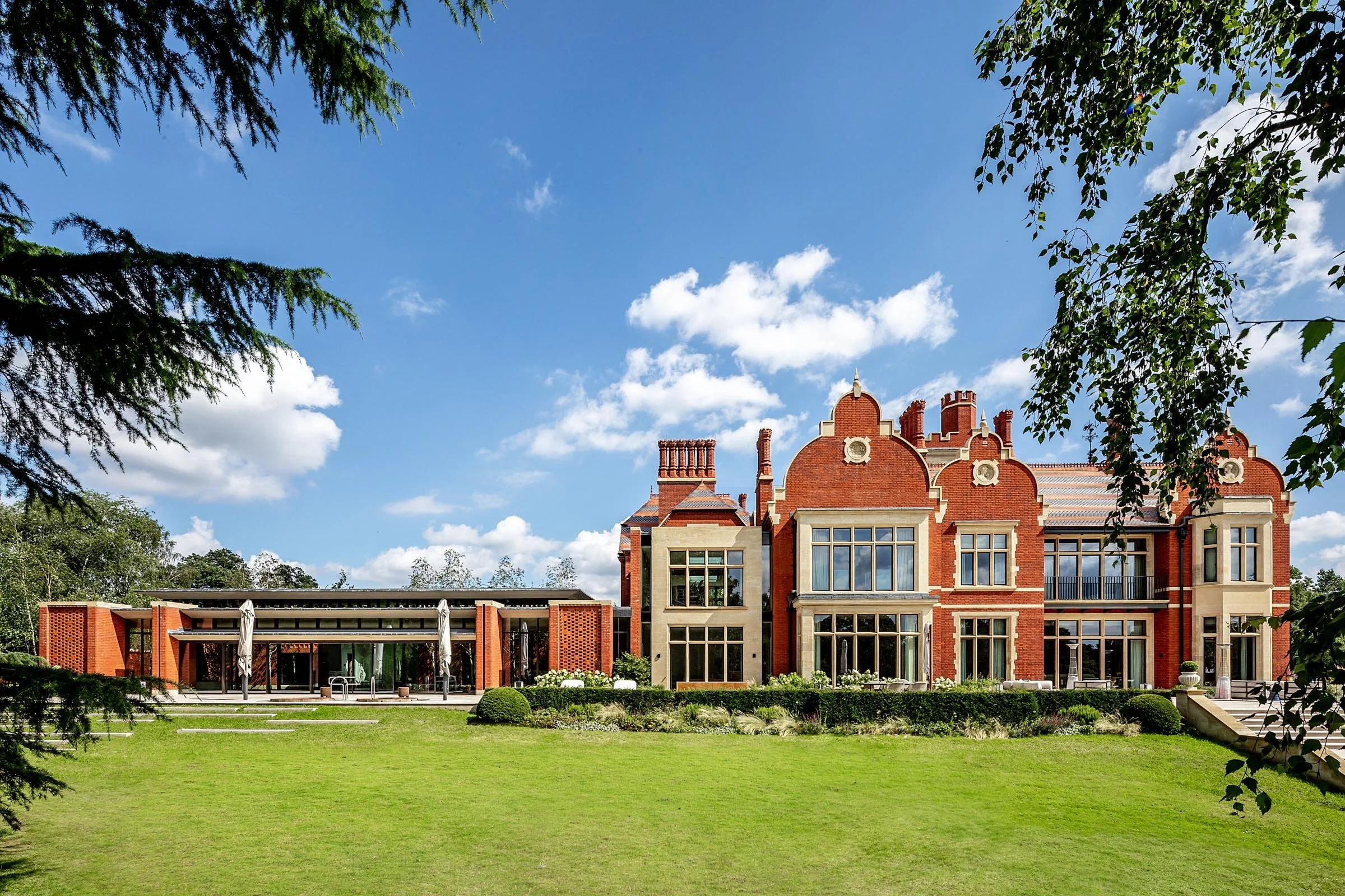 A large historic red brick mansion with multiple chimneys and ornate gables, surrounded by a well-maintained lawn and framed by trees, under a bright blue sky with scattered white clouds.