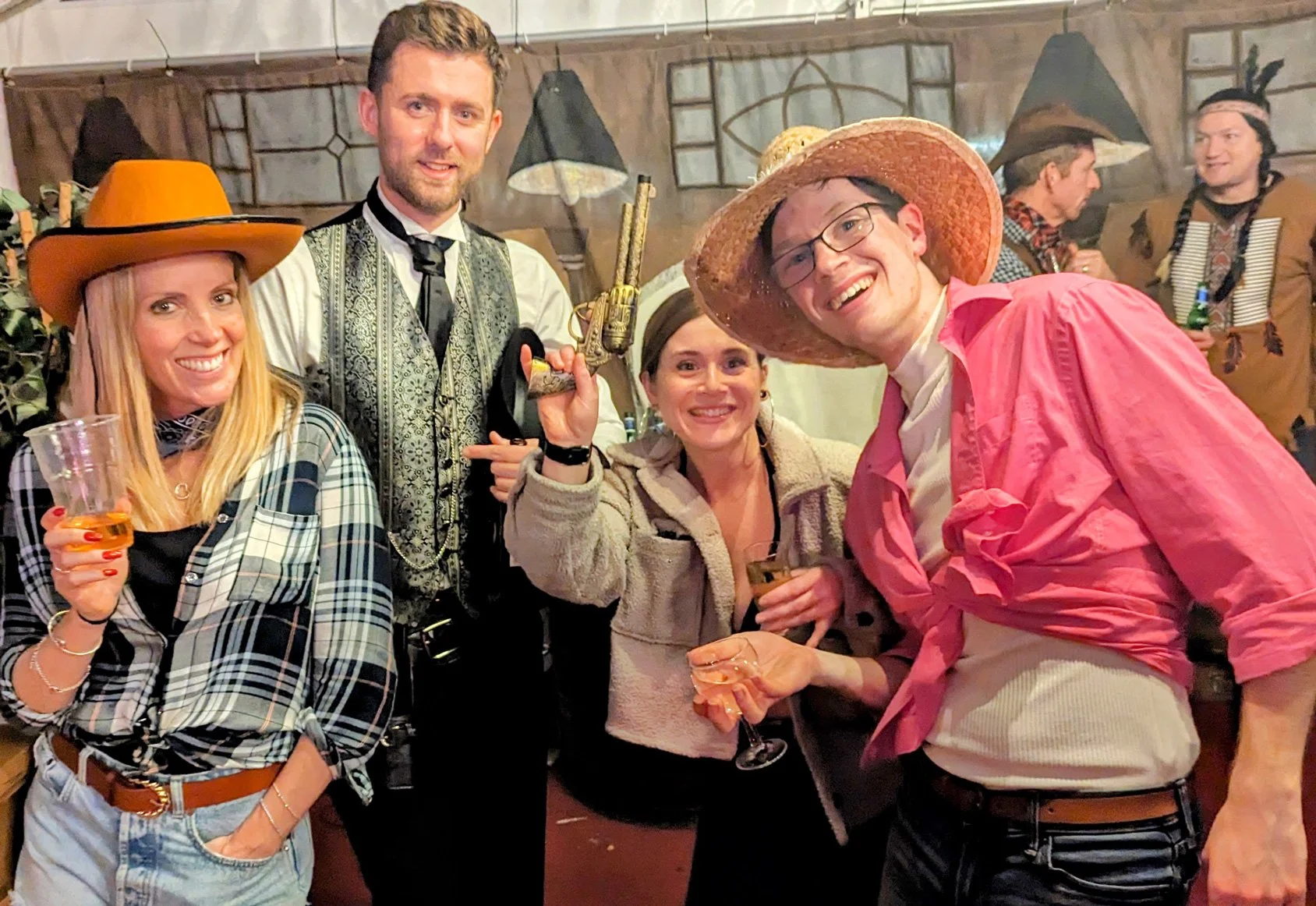 Group of five people at a Western-themed party, dressed in cowboy hats, plaid shirts, and costumes, holding drinks and smiling.