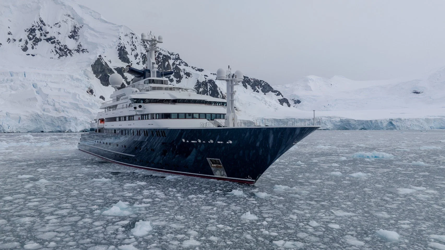 A large luxury yacht sailing through icy Arctic waters with snow-covered mountains in the background.