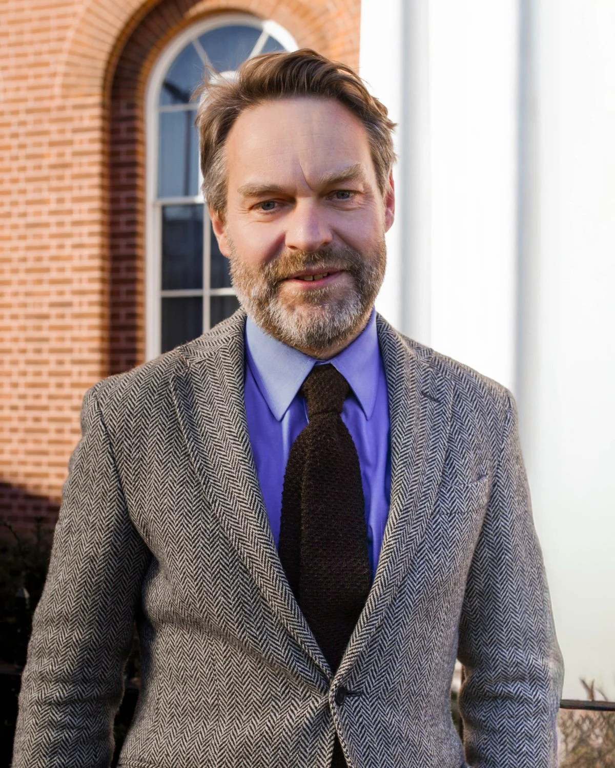 A smiling man with short brown hair wearing a light-colored button-up shirt standing against a blue background.
