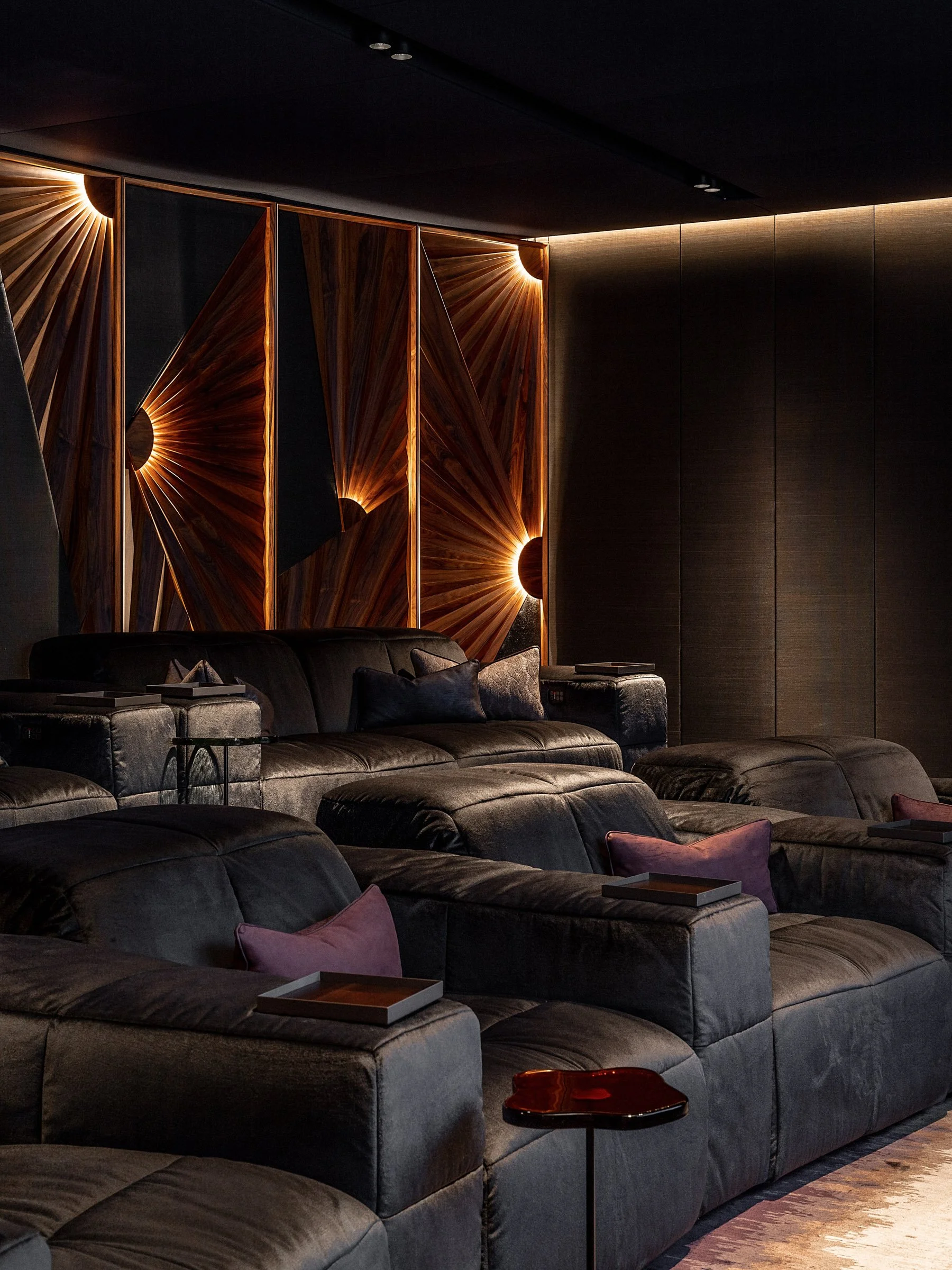 Dimly lit modern living room with black leather couches, purple pillows, and decorative wood wall art with backlighting.