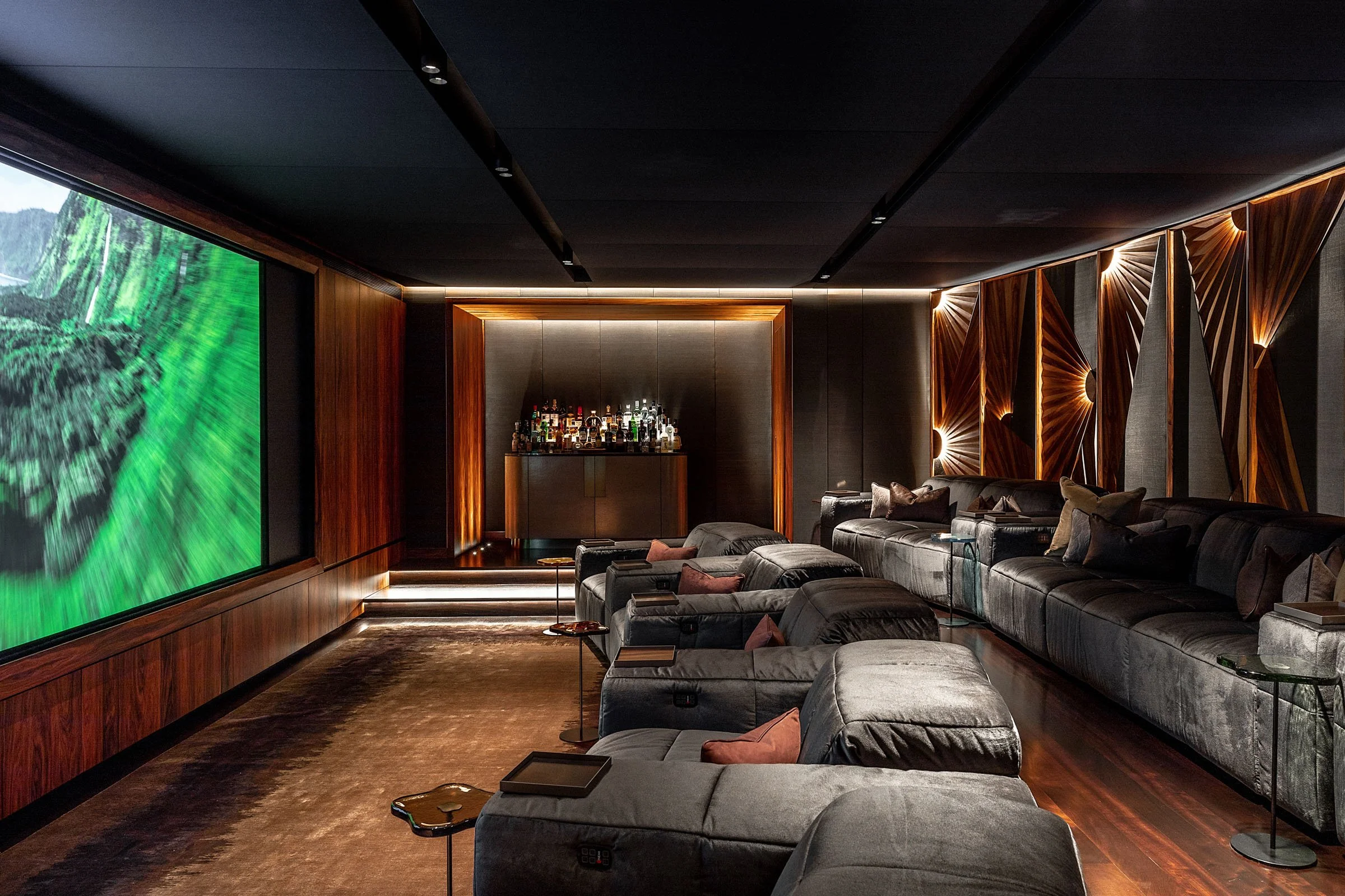 Luxurious home theater room with large screen, plush gray seating, ambient lighting, and a bar area at the back.
