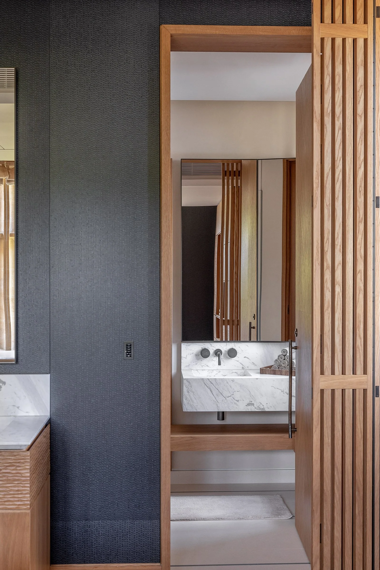 View through a wooden door to a bathroom sink with marble countertop and rolled towels, surrounded by wood accents and a mirror.