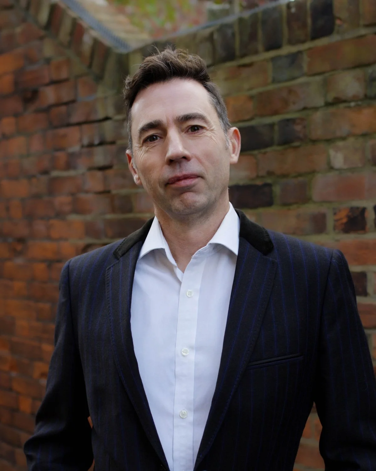 A man with short dark hair, light skin, wearing a salmon-colored collared shirt and a patterned inner shirt, standing against a blue background.