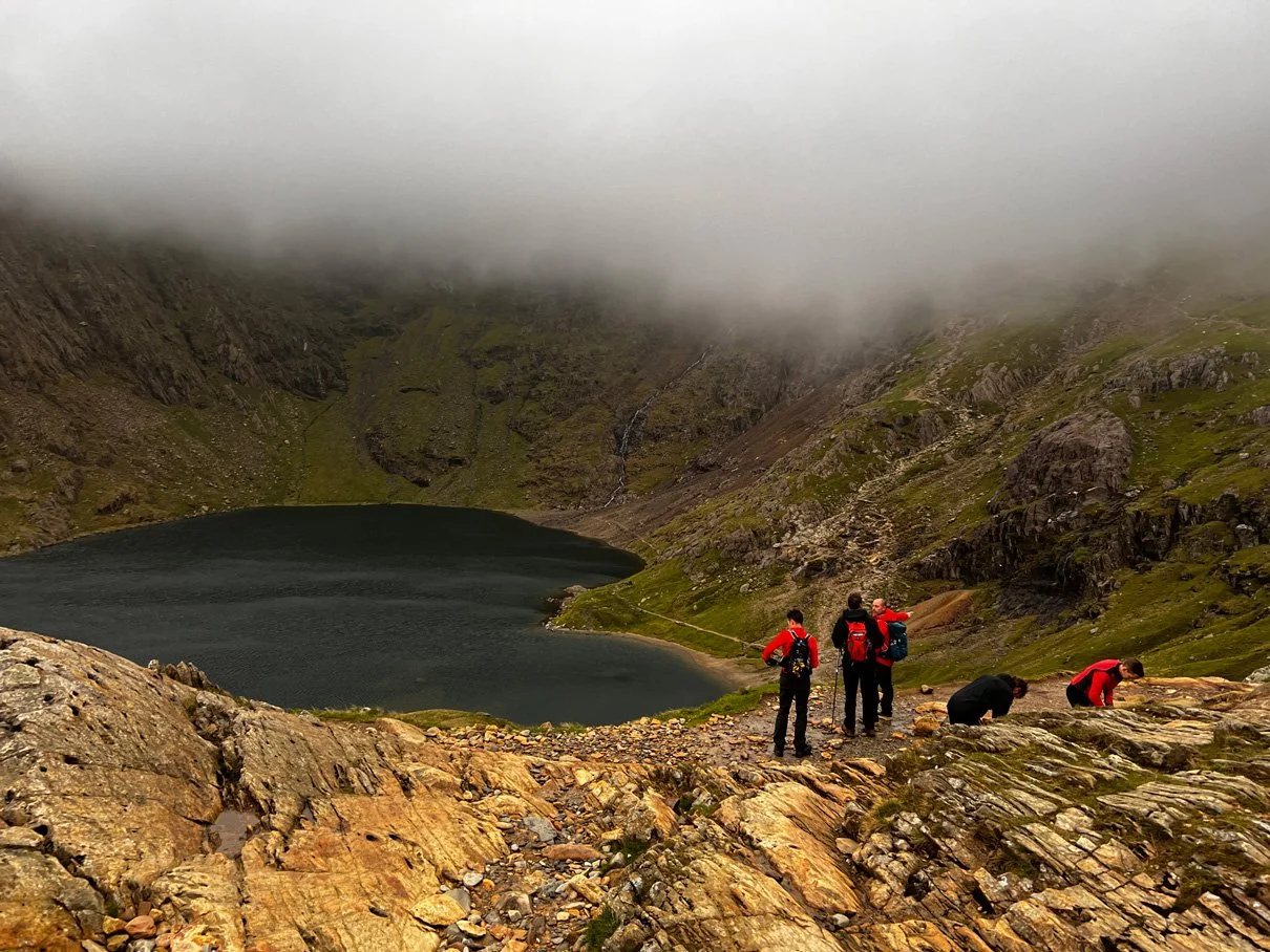 Four hikers in red and black jackets and backpacks on rocky terrain by a mountain lake, with green slopes and fog-covered mountains in the background.