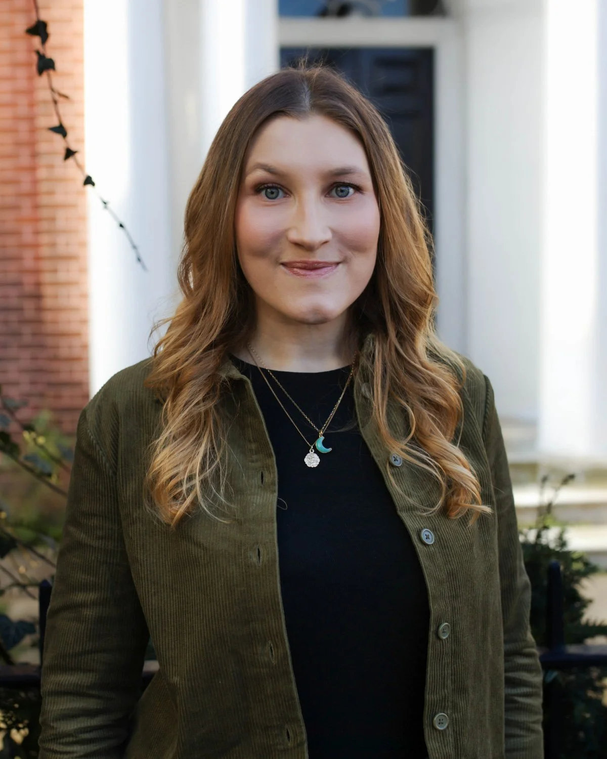 A woman with long wavy red hair, blue eyes, and wearing a black top, posing against a plain white wall.