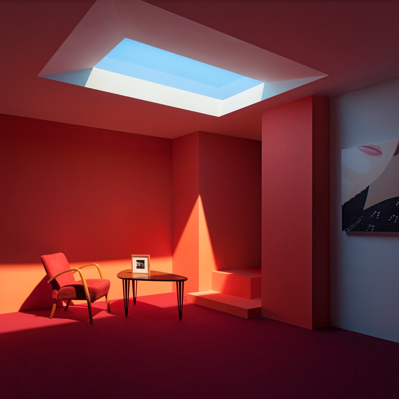 A room with pink walls and a pink carpet, illuminated light coming through a rectangular fake Coelux skylight in the ceiling. There is a pink armchair, a small black side table with a framed photo, and a staircase leading to a hidden area.