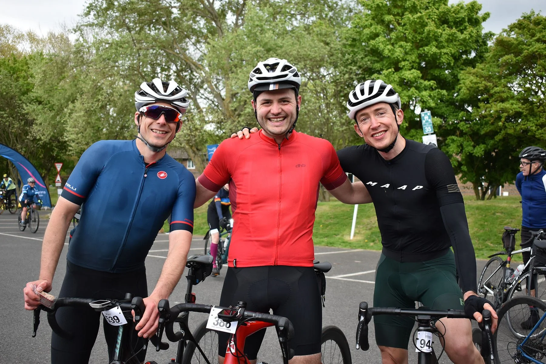 Three male cyclists in racing gear and helmets smiling at the camera, with other cyclists and trees in the background.