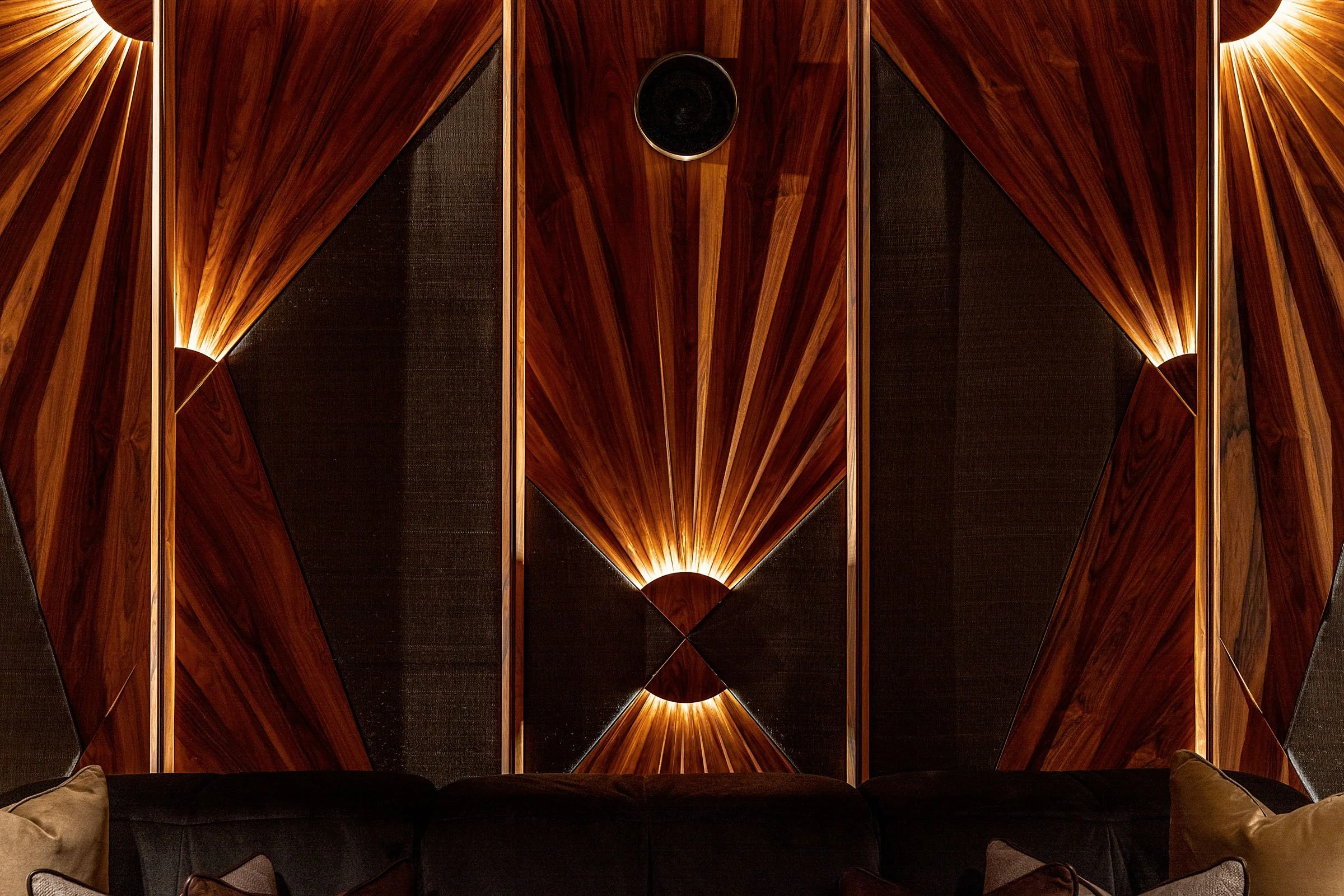 Close-up of a wooden wall with geometric patterns and lighting, with a black circular speaker at the top center, and part of a dark sofa with beige pillows visible at the bottom.