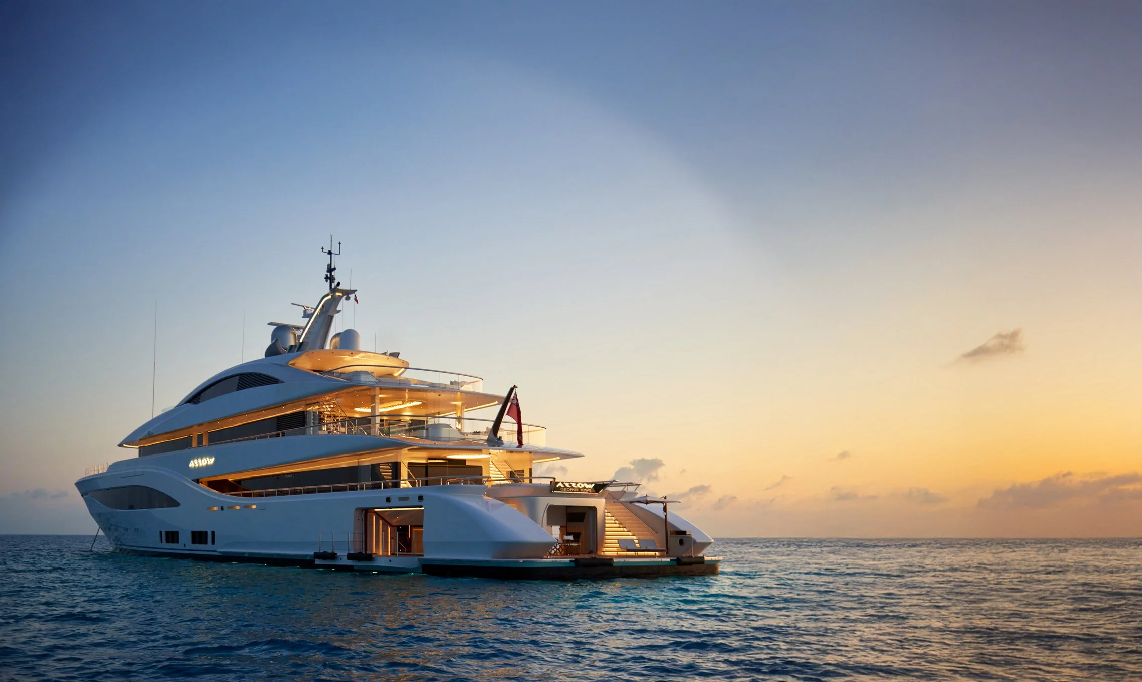 A luxurious white yacht floating on calm ocean waters at sunset, with the sky displaying shades of orange and blue.