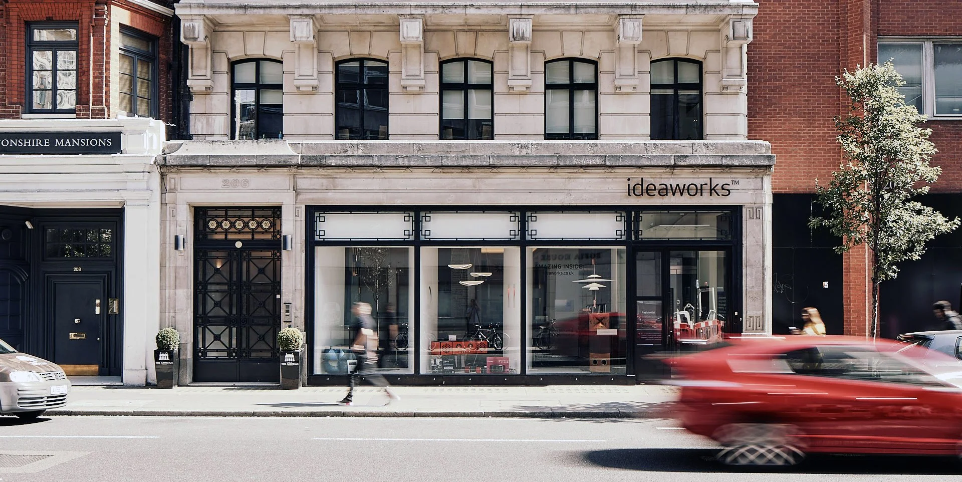 Street view of a commercial building with the sign 'ideaworks,' showing large glass windows inside a modern furniture or decor store, with pedestrians and a passing red car on the street.