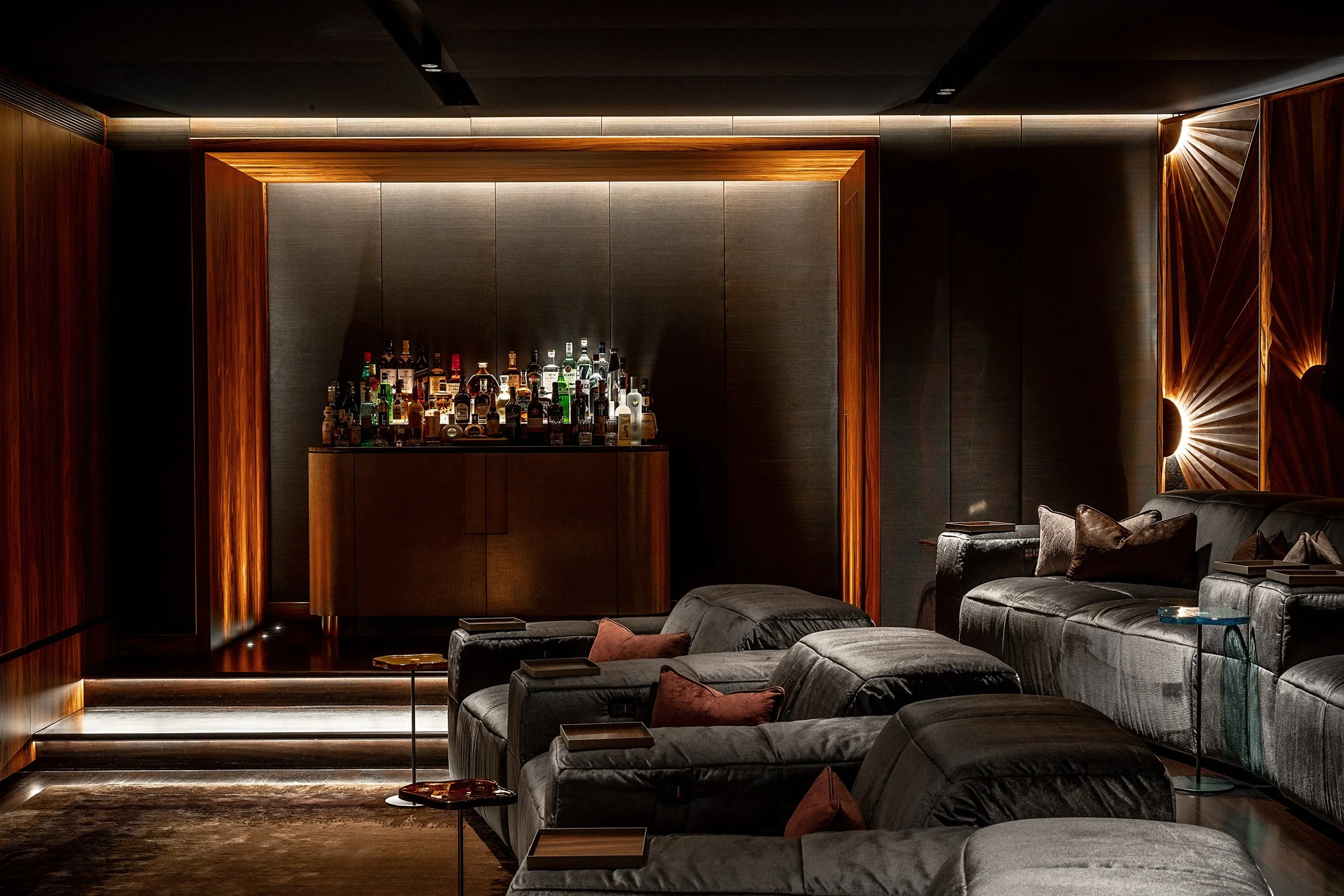 A luxurious lounge with dark wooden walls, plush gray velvet seating with pillows, a bar area with various bottles, and ambient lighting accents.