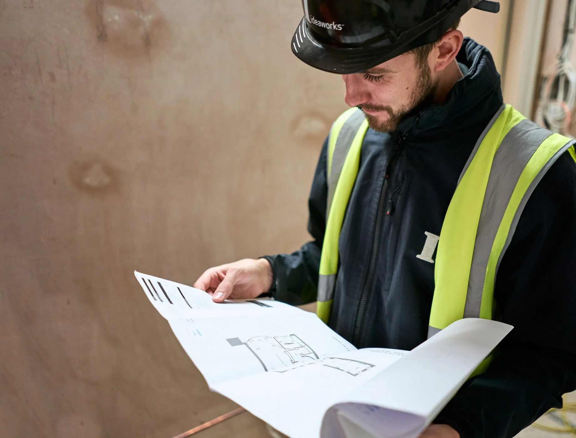 A construction worker wearing a black helmet and a high-visibility safety vest reads a blueprint or construction plan.
