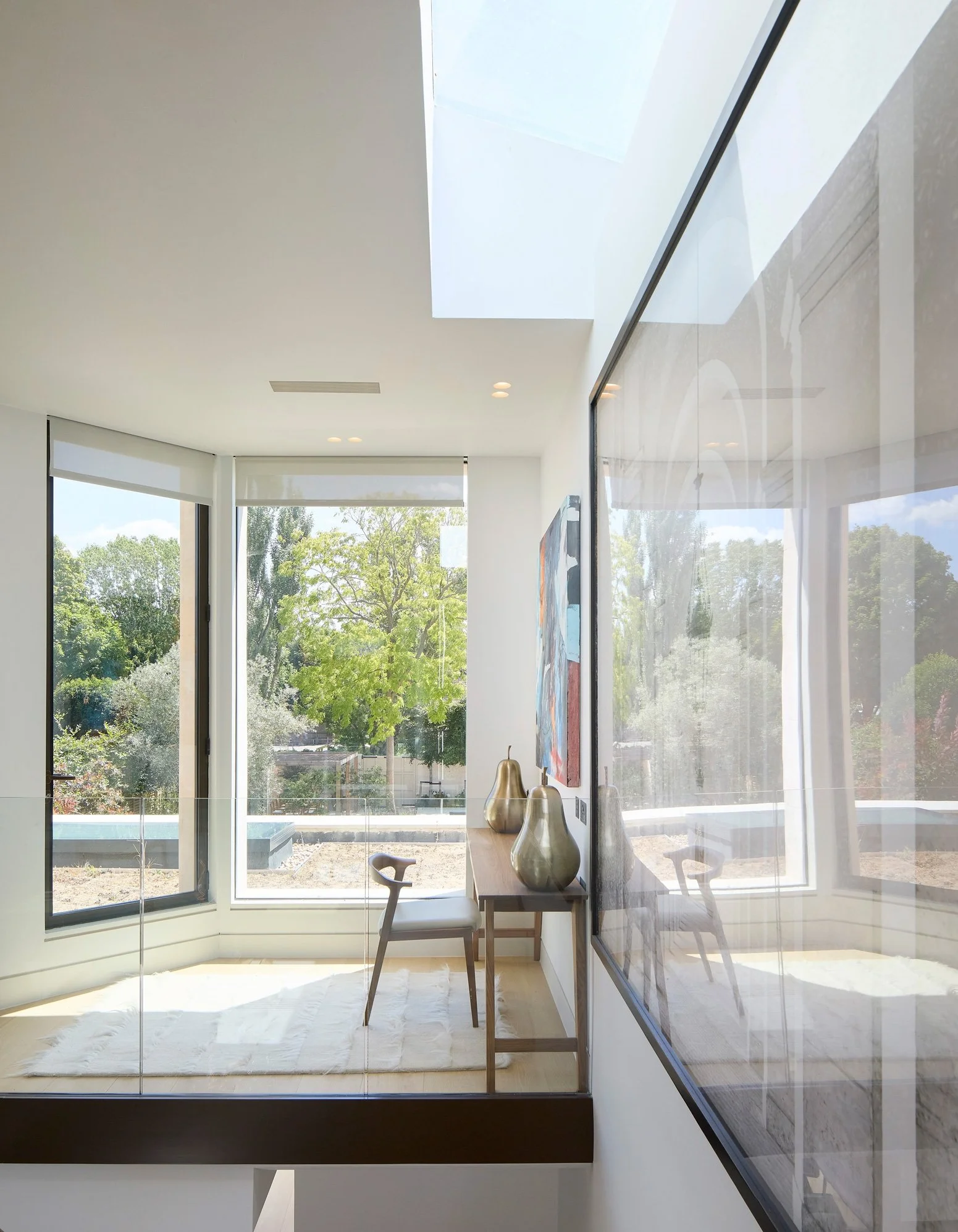 Bright modern interior room with large windows showing greenery outside, a wooden desk with three gold vases, a black and white chair, and abstract artwork on the wall.