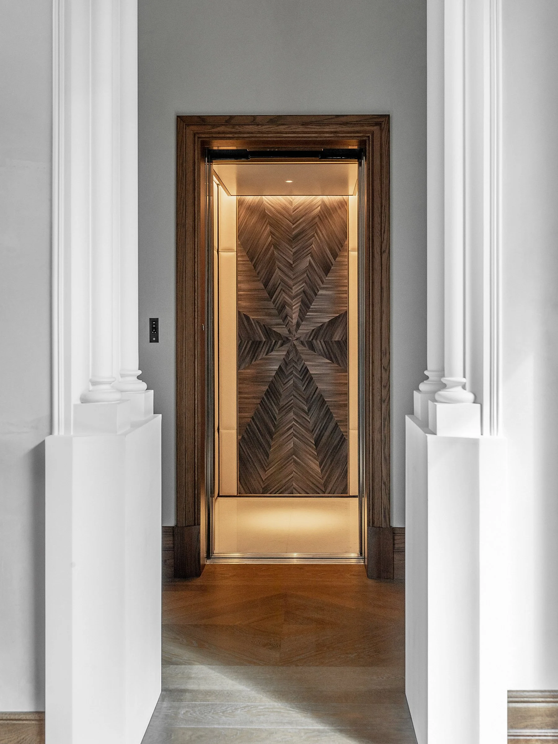 Emotionally evocative image of a modern design elevator with intricate wood paneling on the door, surrounded by classic white columns and trim, in a stylish interior.