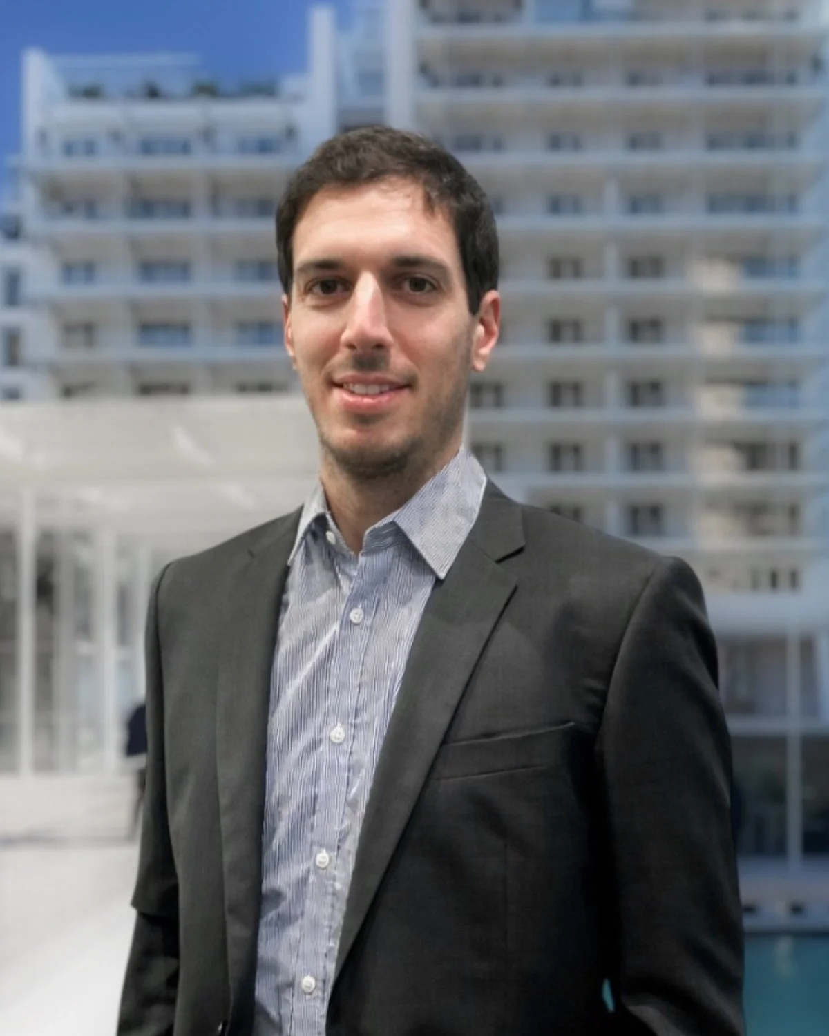 Portrait of a young man with short dark hair, smiling, wearing a black polo shirt, standing against a plain light-colored wall.