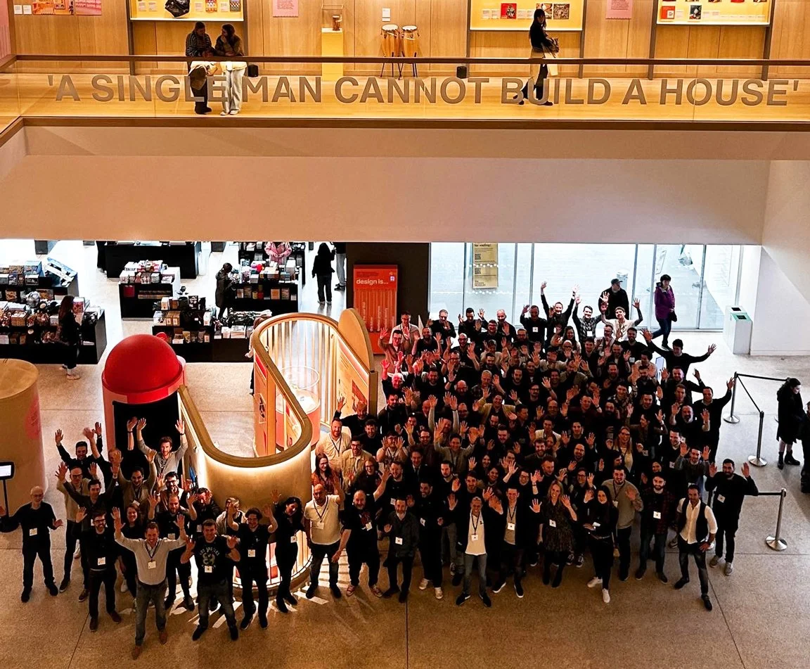Large group of people gathered inside a modern building, smiling and waving at the camera. The building has high ceilings and a second-floor balcony with a sign that reads 'A single man cannot build a house.'
