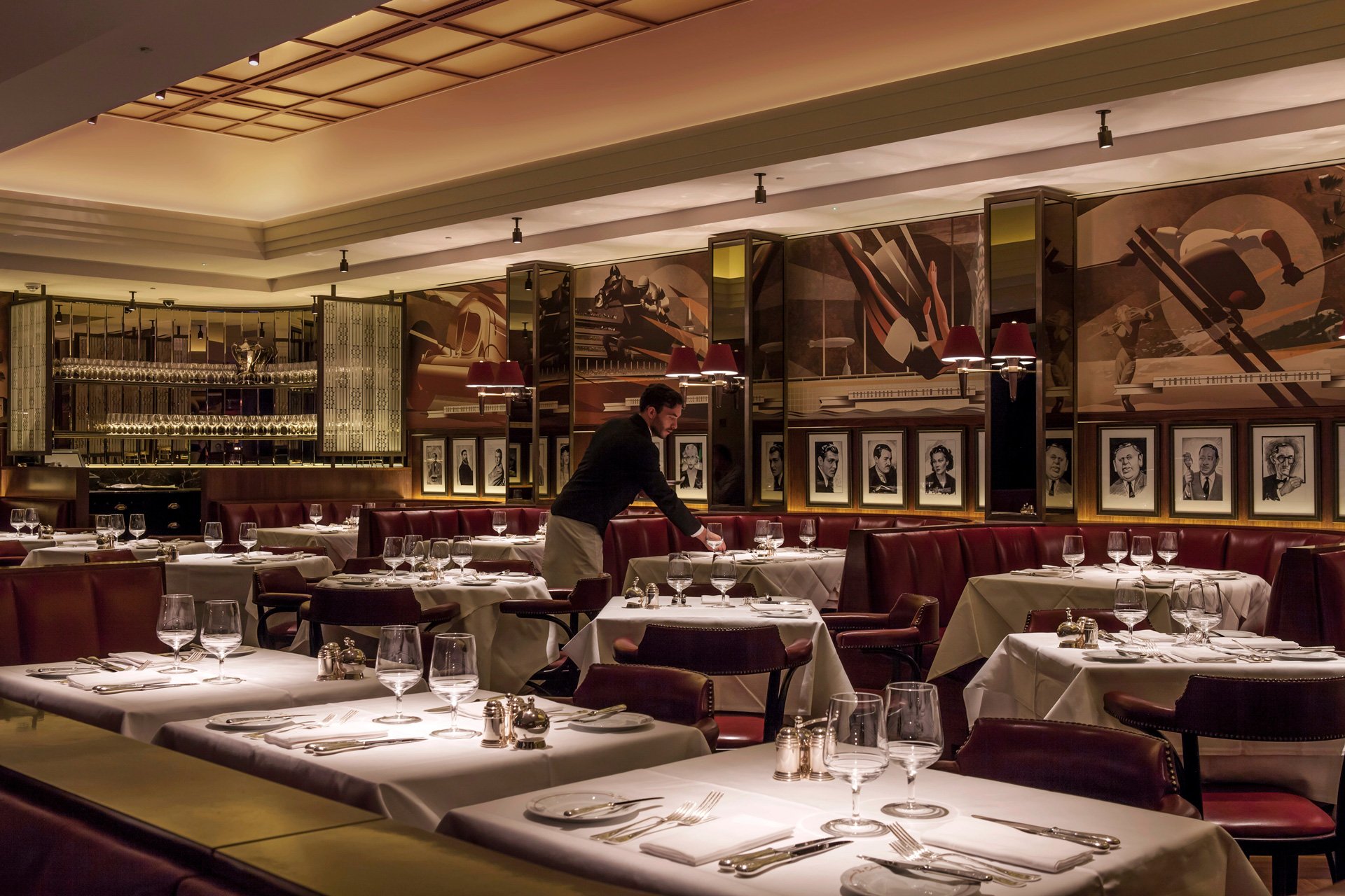 Empty upscale restaurant with tables set for dining, a server arranging a table, and large wall art featuring portraits and musical instruments, warm lighting, and a modern elegant interior.