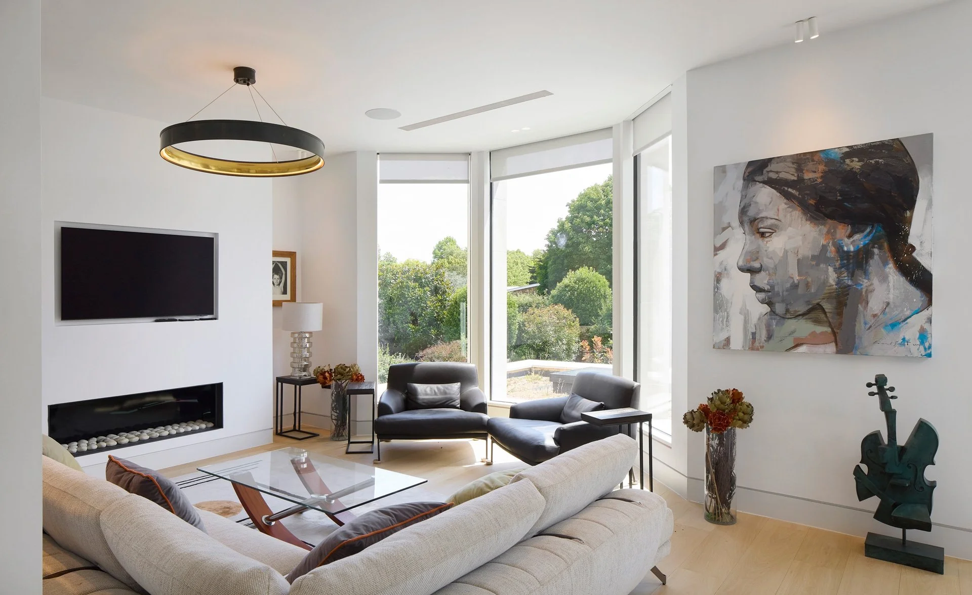 Modern living room with large windows, a beige sectional sofa, black armchairs, a fireplace, a wall-mounted TV, artwork, and decorative items.