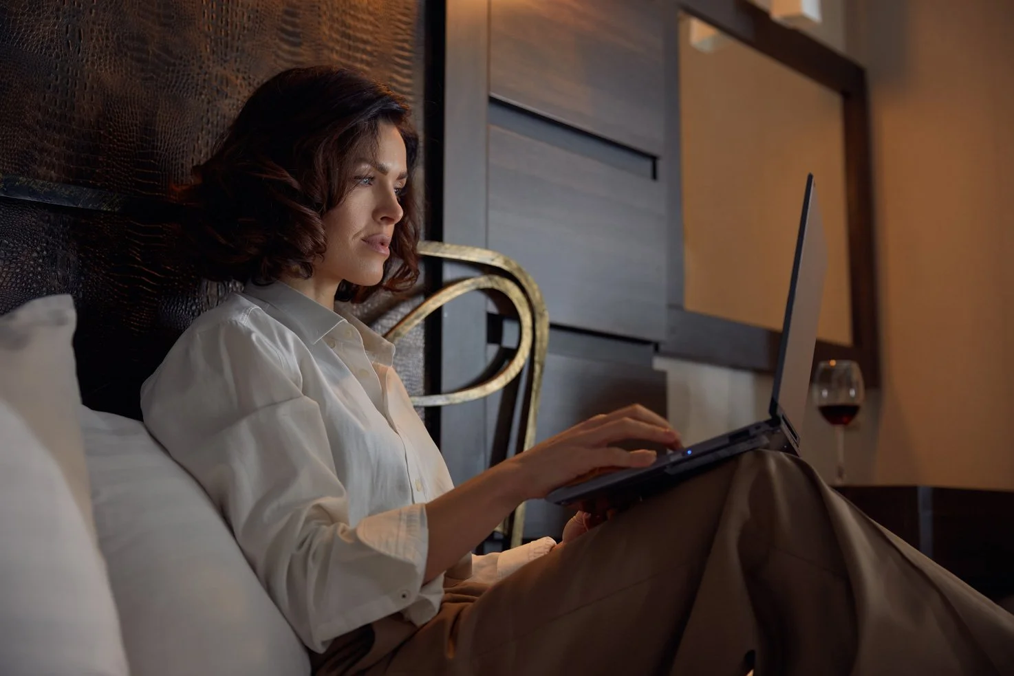 Woman with short brown hair lying on bed using a laptop, with a glass of red wine on nightstand beside her