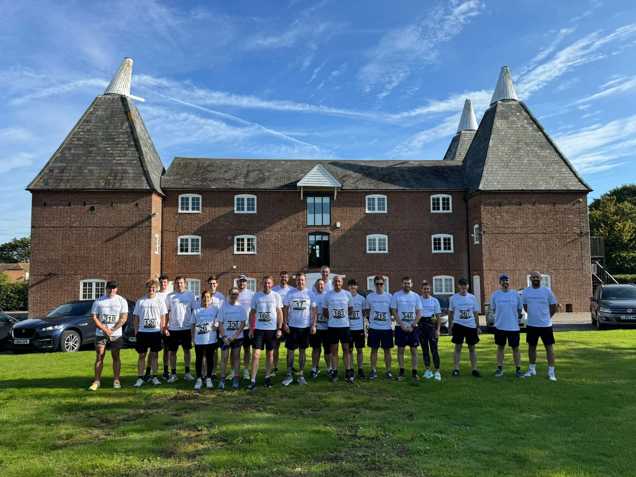 Group of runners in white t-shirts with numbered bibs standing on grass in front of a large brick castle-like building with four tall conical towers and a blue sky with clouds.