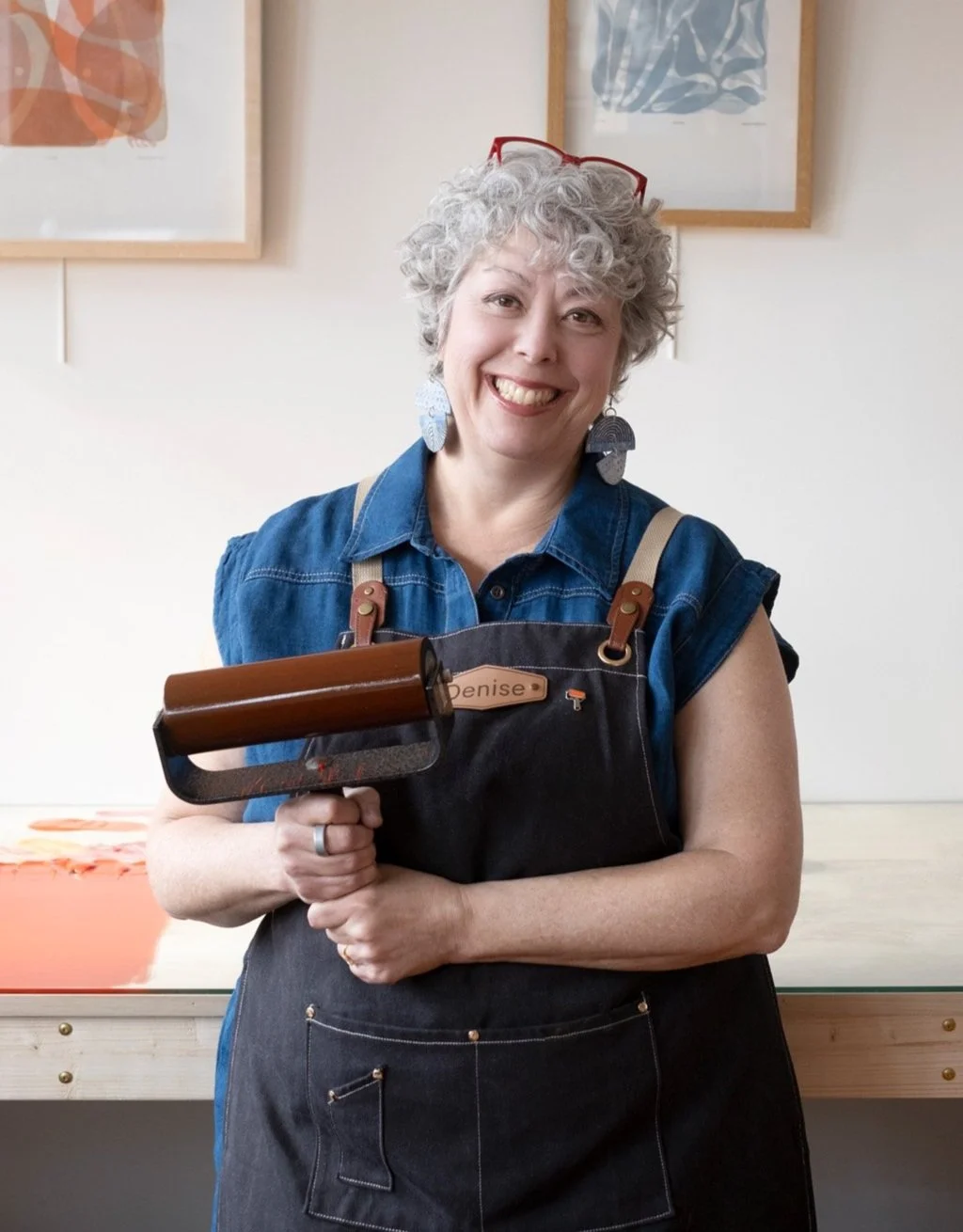A smiling Denise Markbreit in her Asheville, NC printmaking studio wearing a black smock and holding a large inking roller covered in red eco-friendly Akua ink.
