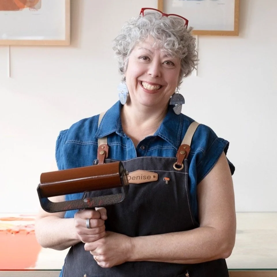 A smiling Denise Markbreit in her Asheville, NC studio clothes holding a large inking roller covered in red environmentally-friendly Akua ink.