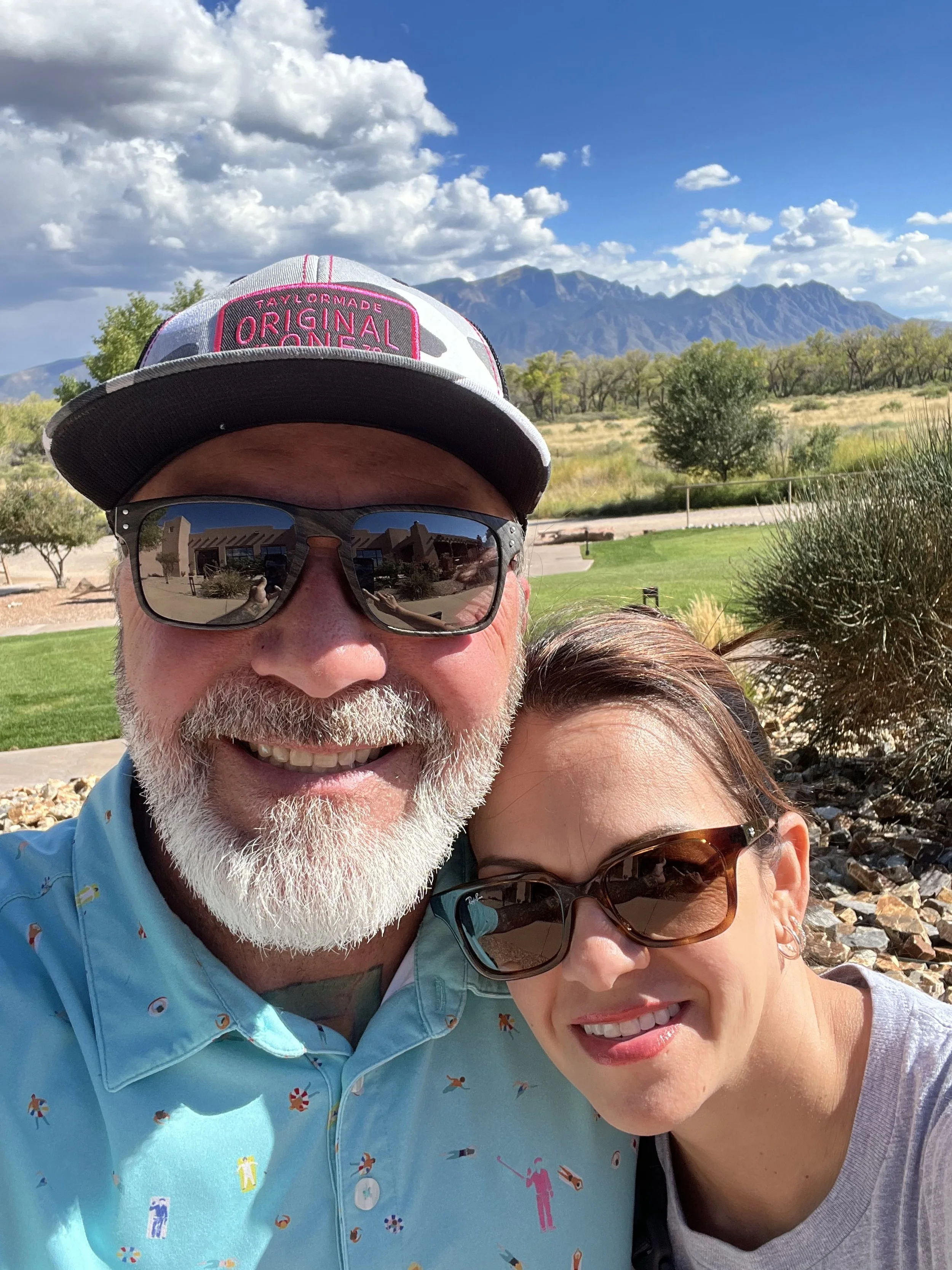 A man and woman taking a selfie outdoors, both wearing sunglasses, with a clear blue sky, mountains, and a green landscape in the background goodlife construction