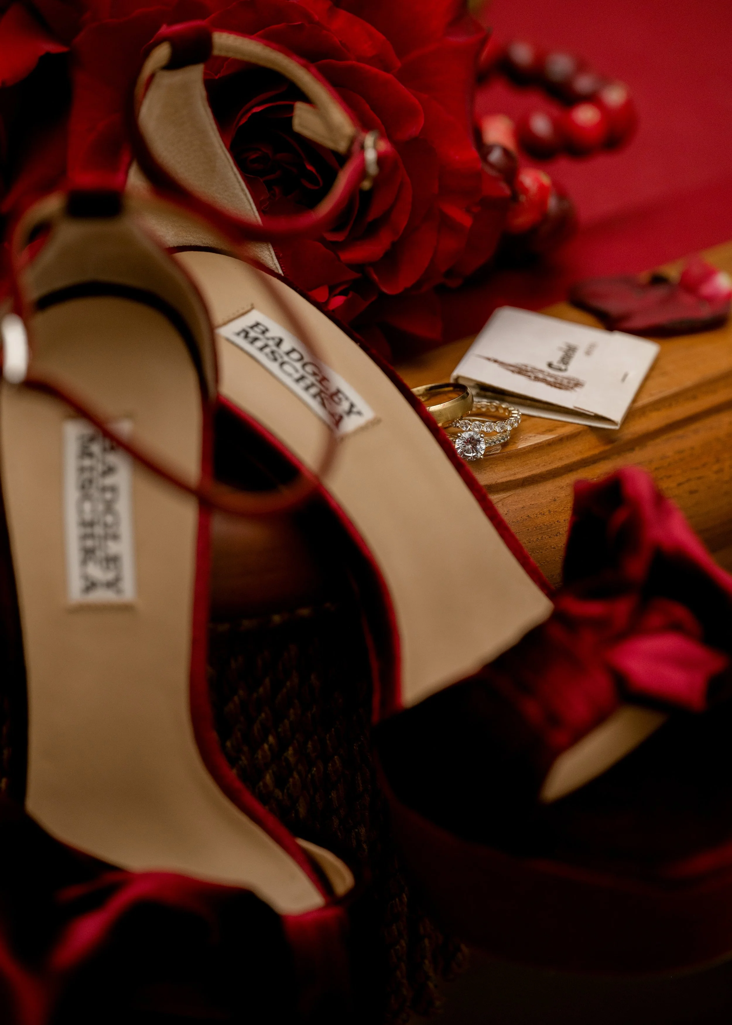 Red high-heeled shoe, red roses, jewelry including a diamond engagement ring, and a small booklet on a wooden surface.