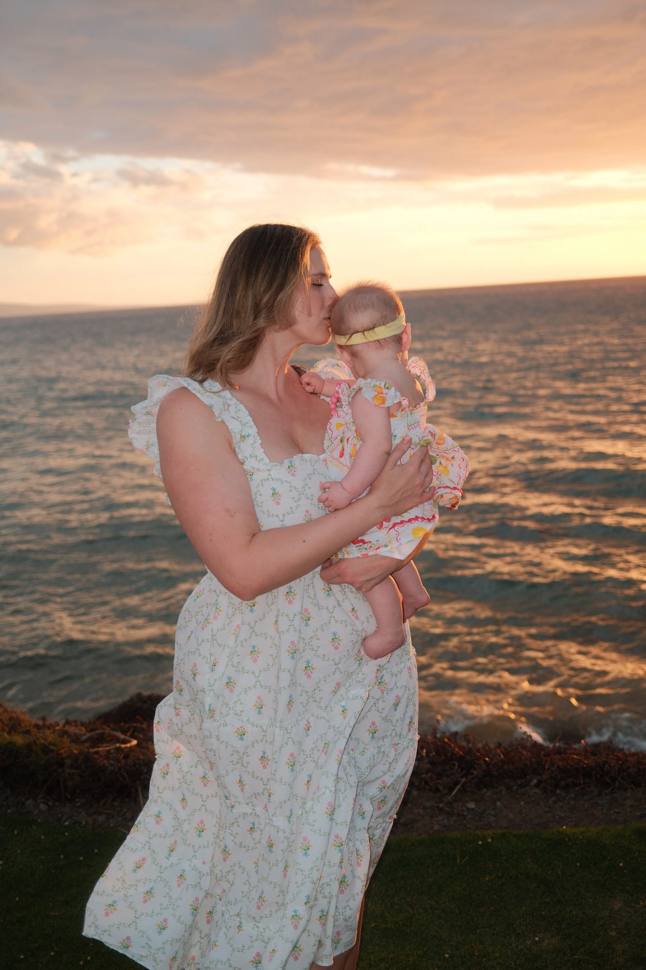 A woman holding a baby by the ocean at sunset, both dressed in white floral dresses, with the woman kissing the baby's forehead.