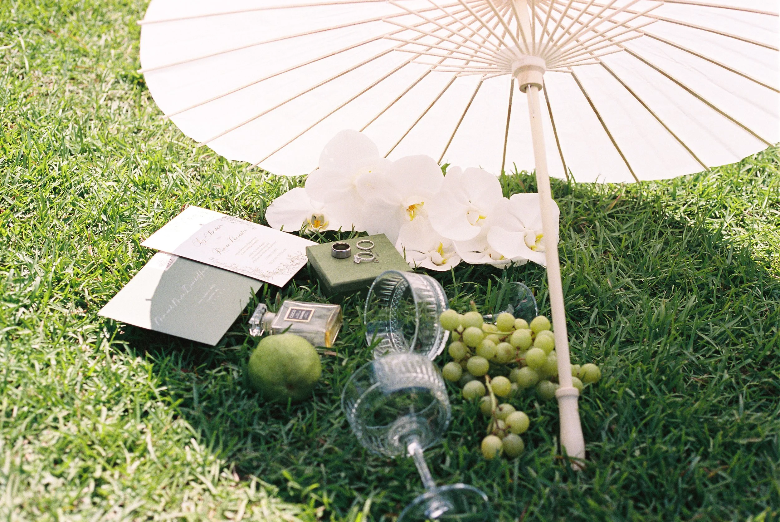A white paper parasol resting on the grass surrounded by white orchids, green grapes, a green lime, perfume bottles, rings, and a wedding invitation, suggesting a romantic outdoor setting.