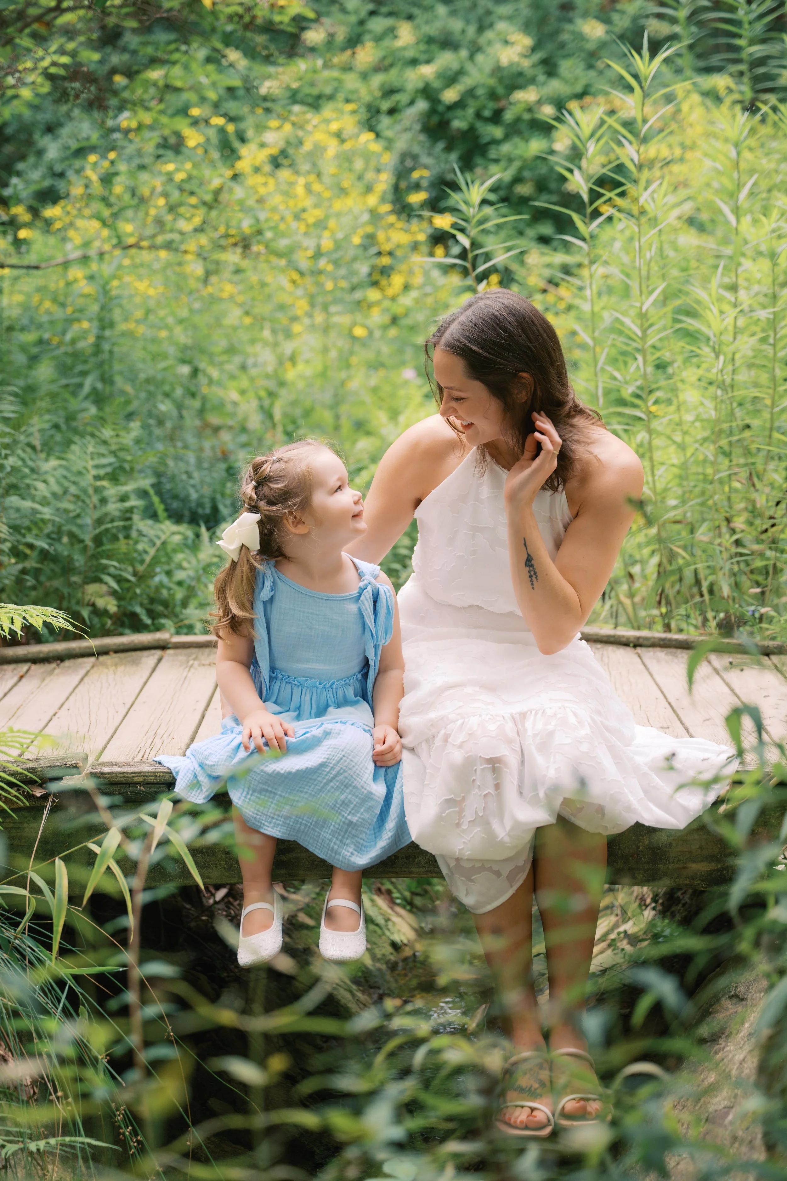 A woman and a young girl sitting on a wooden bridge over water, surrounded by green plants and trees, sharing a joyful moment.