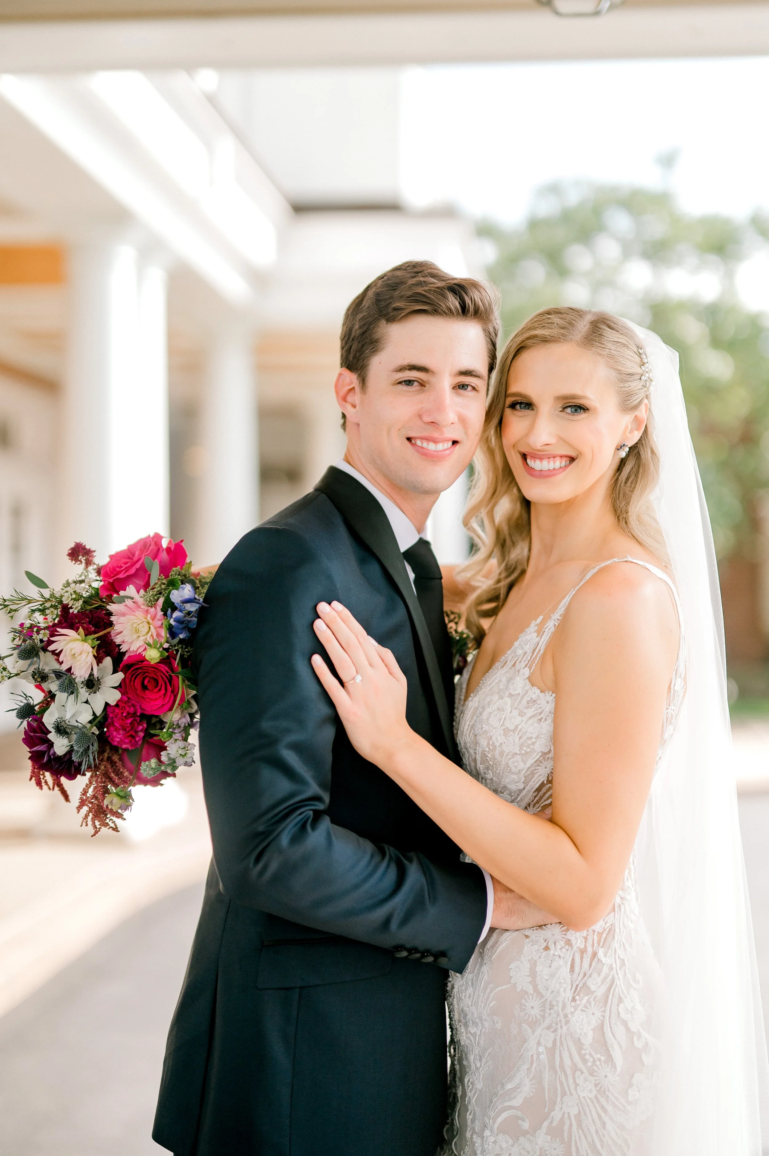 A newlywed couple dressed in wedding attire, standing close together, smiling, and holding each other. The groom is in a black tuxedo, and the bride is in a lace wedding dress with a veil, holding a colorful bouquet of flowers.
