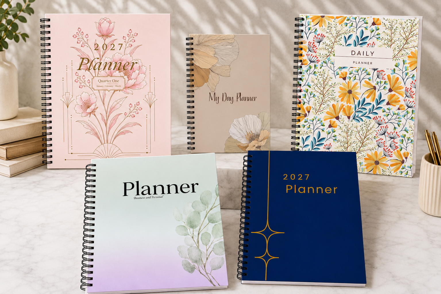 Five spiral-bound 2027 dated and undated planners with floral and botanical cover designs are arranged on a marble surface, with a potted plant and stacked books in the background.