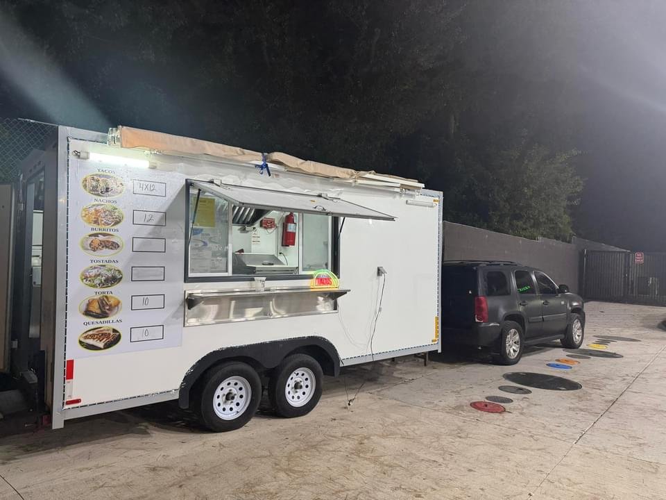 White food truck with menu pictures and prices on the side, parked in a lot with a black vehicle nearby, night time setting.