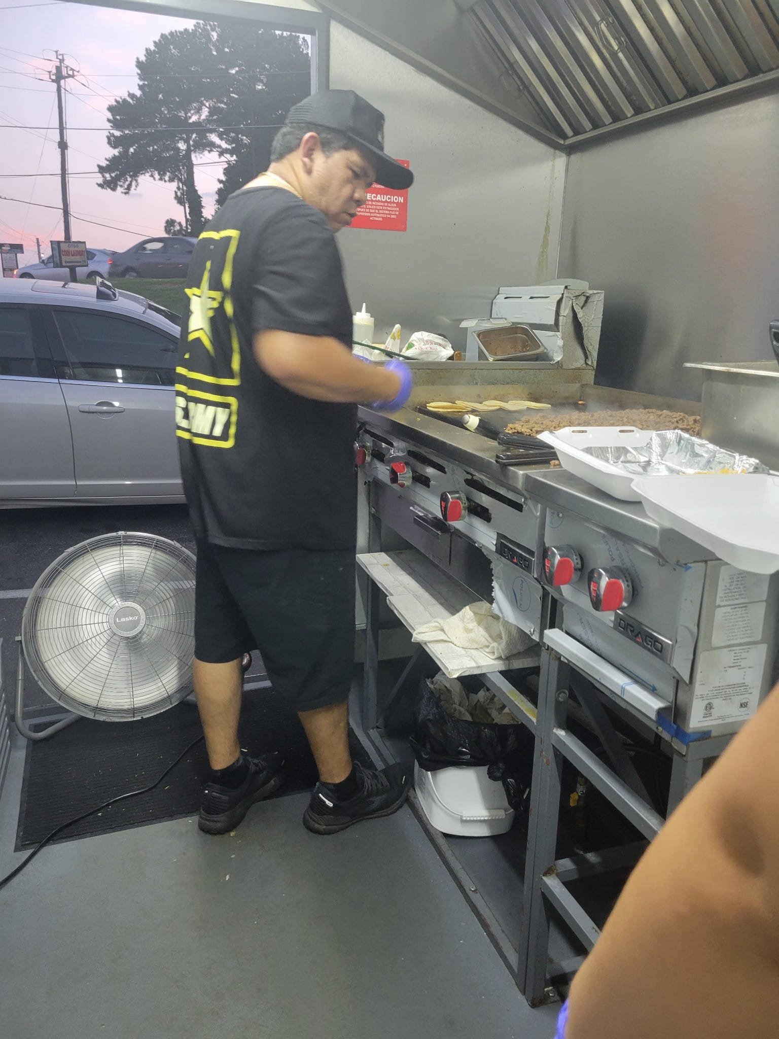 A man cooking on a flat-top grill inside a restaurant kitchen, with a parked car visible outside through a window.