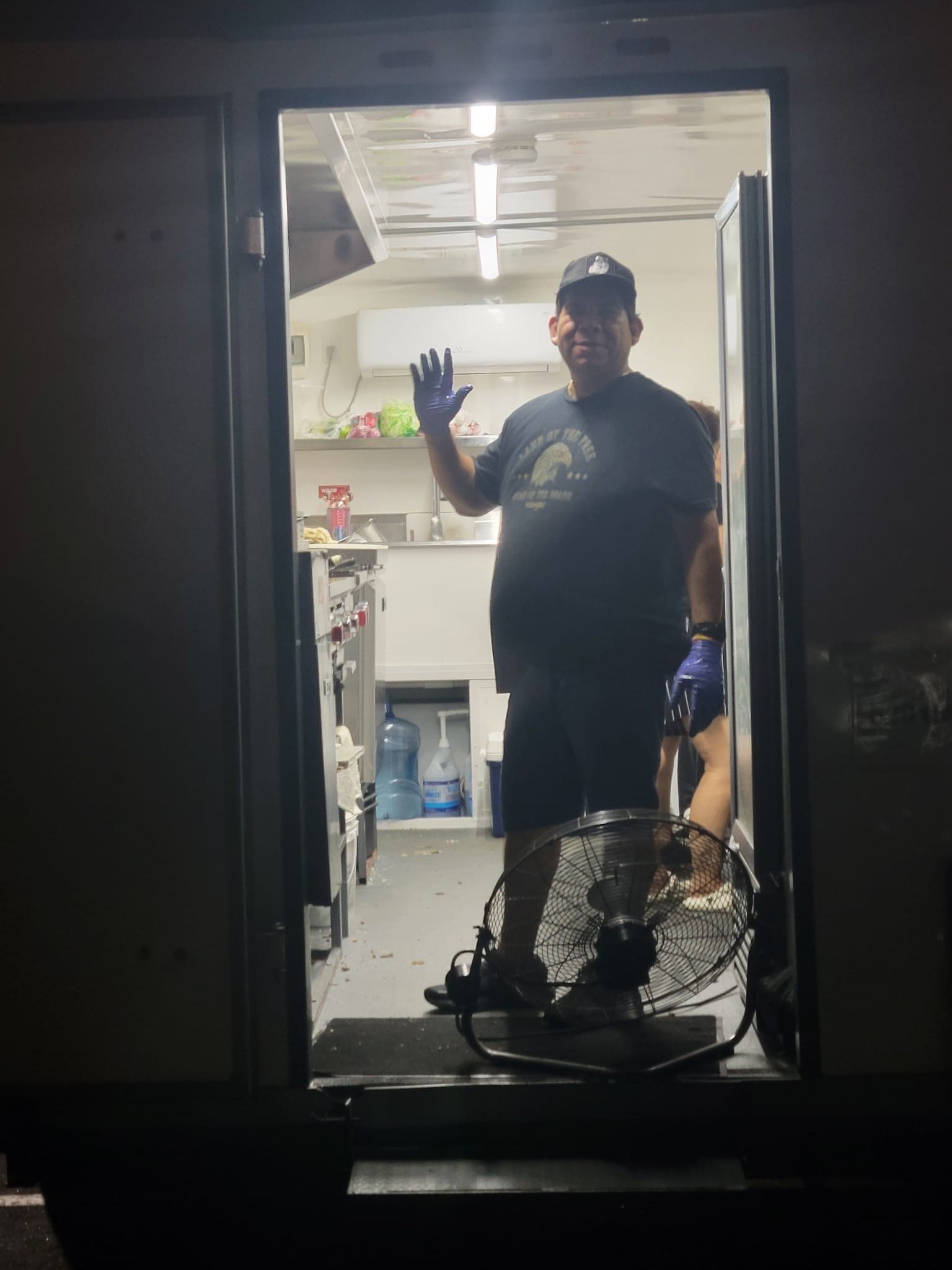 Man wearing a baseball cap, dark t-shirt, and gloves, standing inside a small kitchen and smiling at the camera while waving. The photo is taken through an open window from outside.