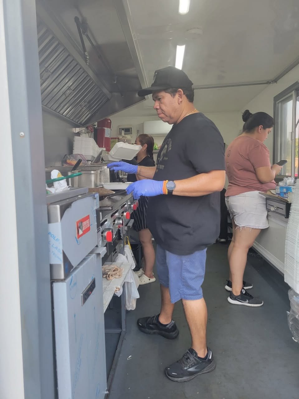 Man in black t-shirt, blue shorts, gray sneakers, and black cap cooking in a food truck kitchen, with two women in the background, one using a phone and the other preparing food.
