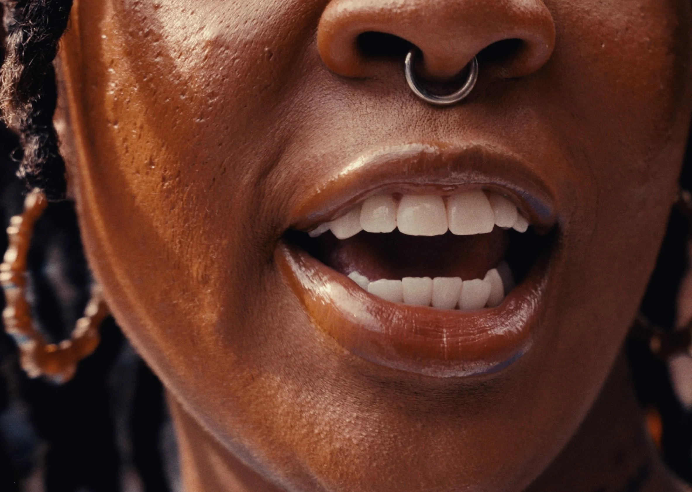 Close-up of a person's face showing a wide smile with white teeth, a septum piercing, and textured skin, with dark braided hair visible on the side.