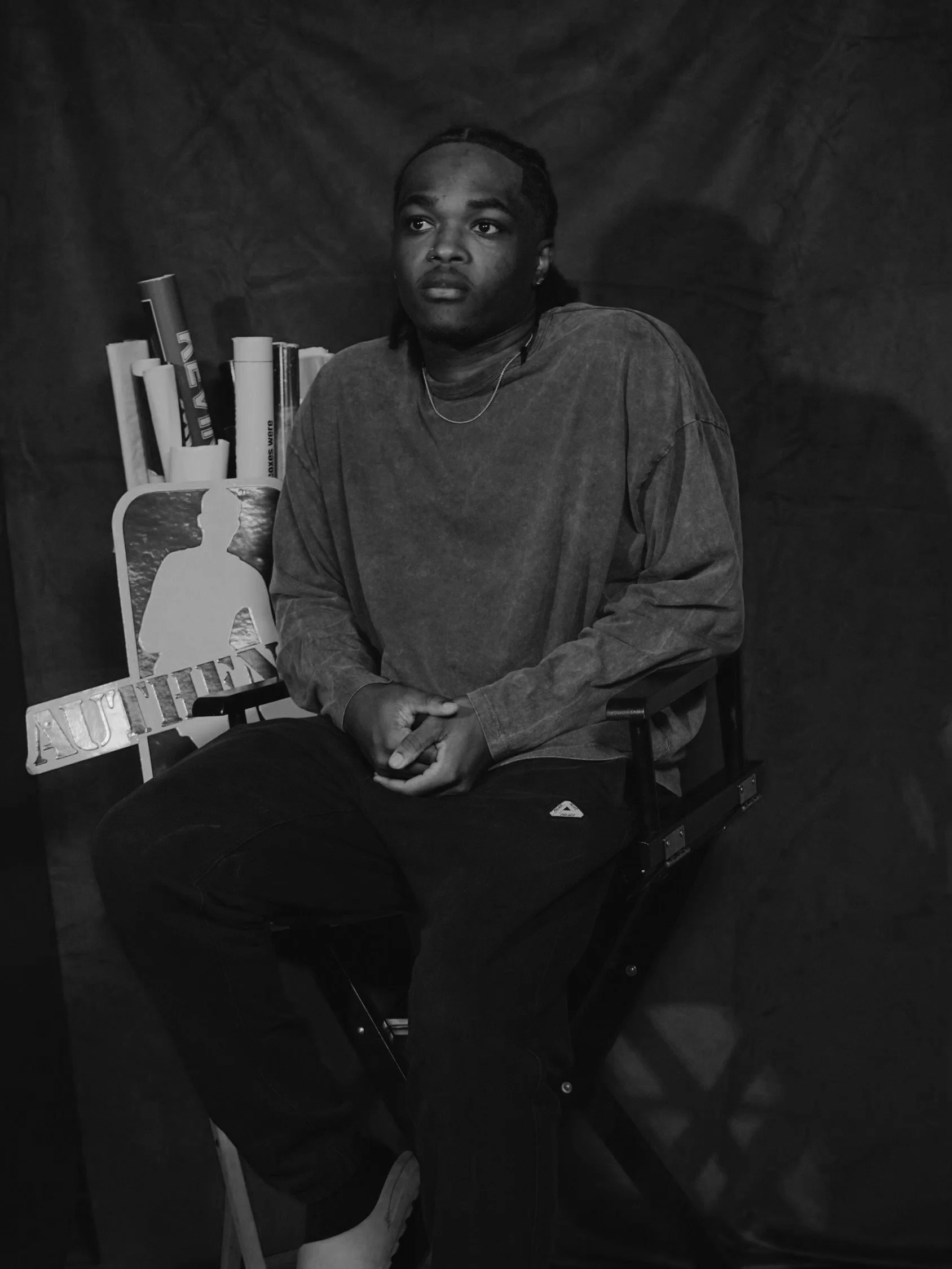 A man sitting on a director's chair in front of a dark background with books and an 'AUTH' sign behind him.