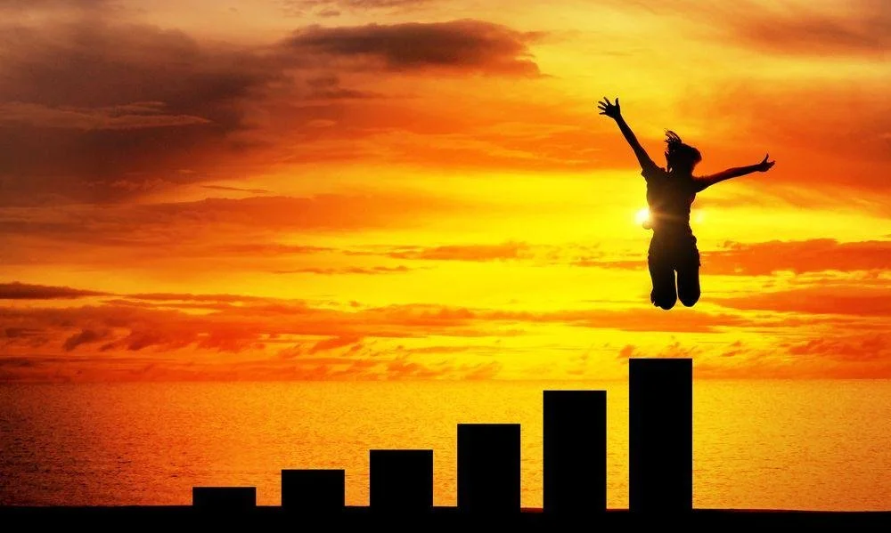 Silhouette of a person jumping in front of a sunset over the ocean, with an upward trending bar graph in the foreground.