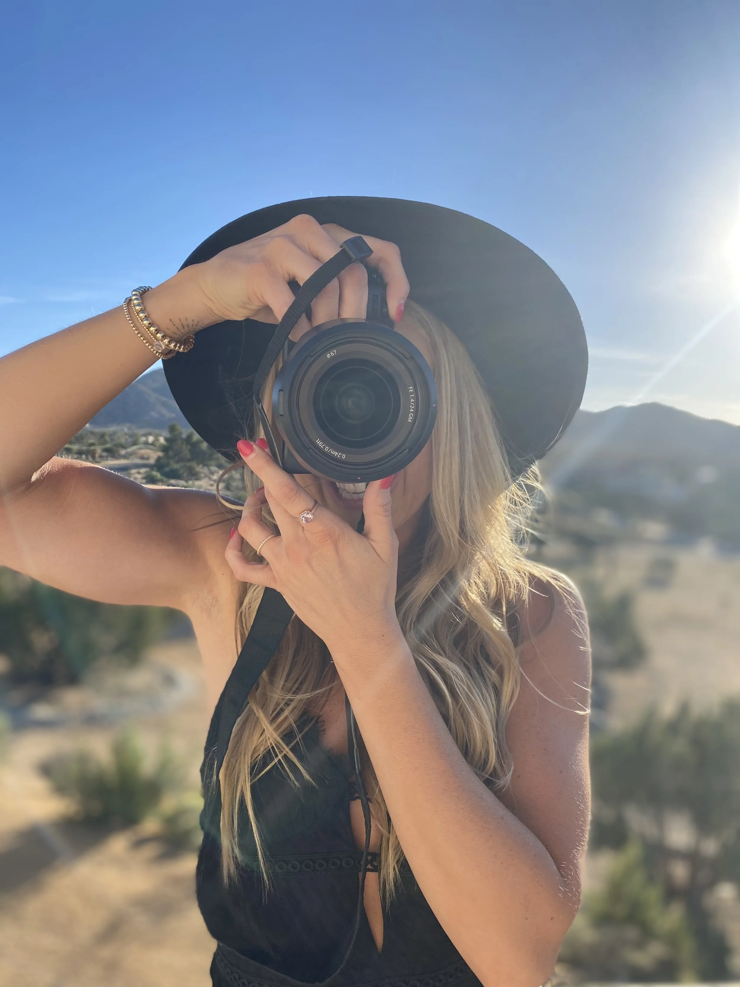 A woman wearing a wide-brimmed black hat and a black sleeveless dress is taking a selfie with a camera, outdoors on a sunny day with mountains and clear blue sky in the background.